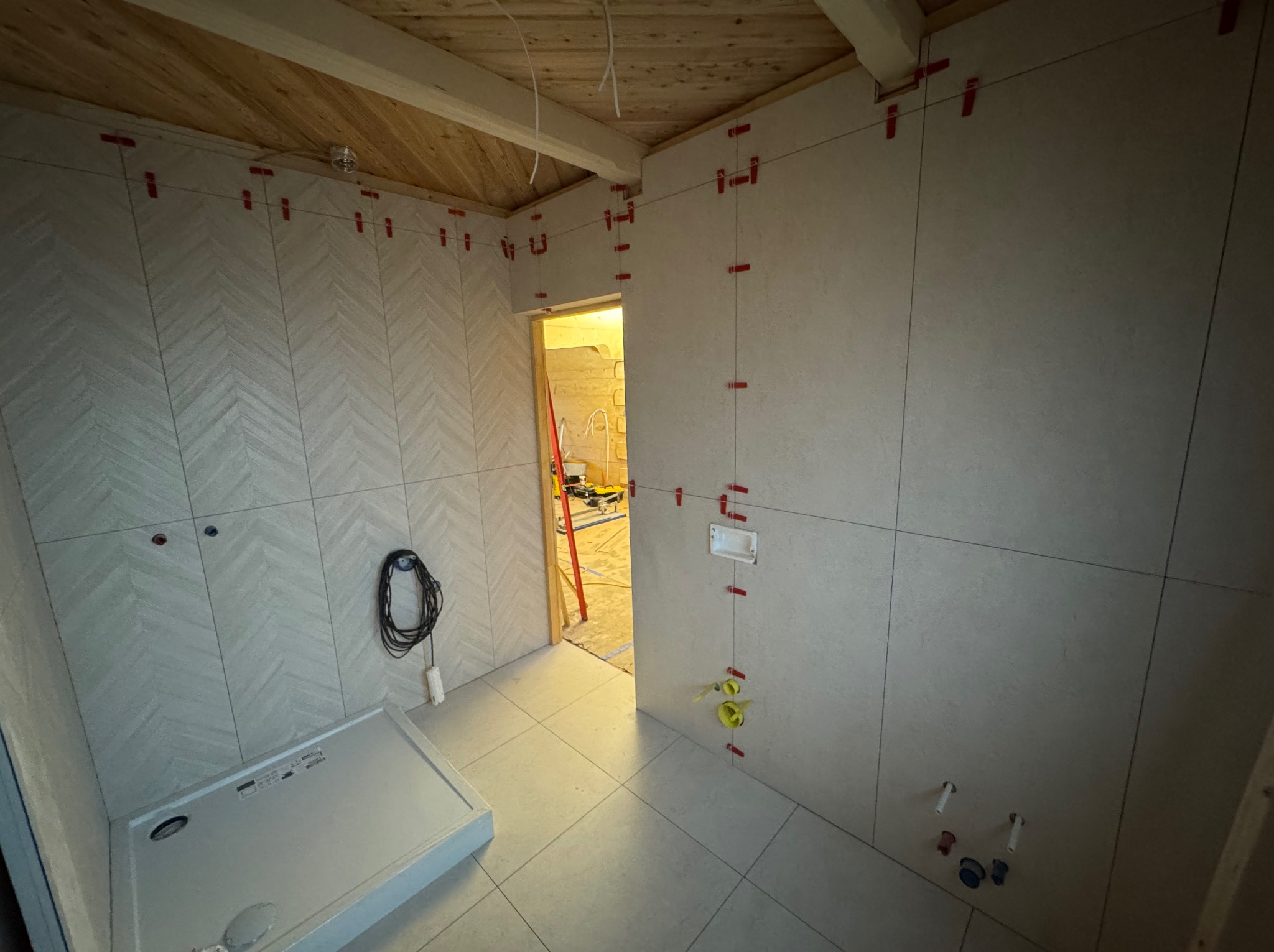 Wnętrze łazienki w trakcie prac wykończeniowych: ułożone płytki w jodełkę na ścianie, brodzik, widoczne elementy instalacji. Czerwone kliny poziomujące płytki na ścianach.