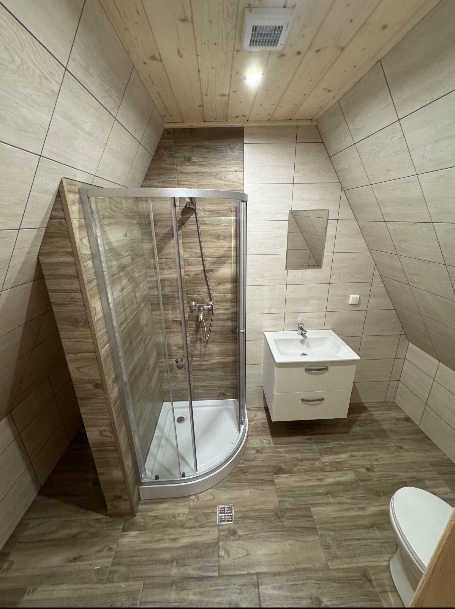 Wykończona łazienka na poddaszu z kabiną prysznicową, umywalką z szafką i toaletą. Ściany i podłoga wyłożone płytkami imitującymi drewno.