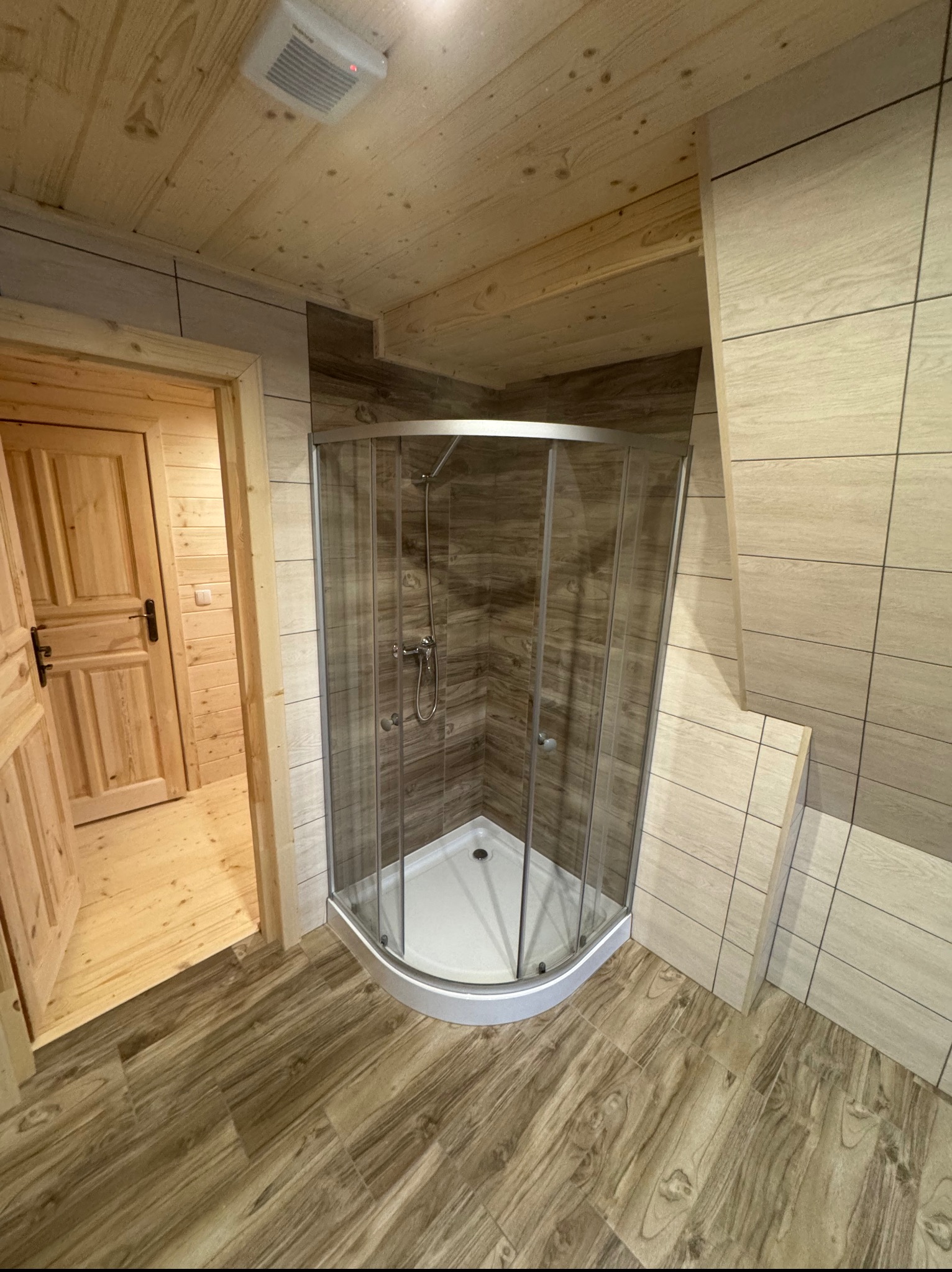 Drewniana łazienka z kabiną prysznicową, wykończona płytkami imitującymi drewno. Widoczny fragment otwartych drzwi i drewniane wykończenie sufitu.