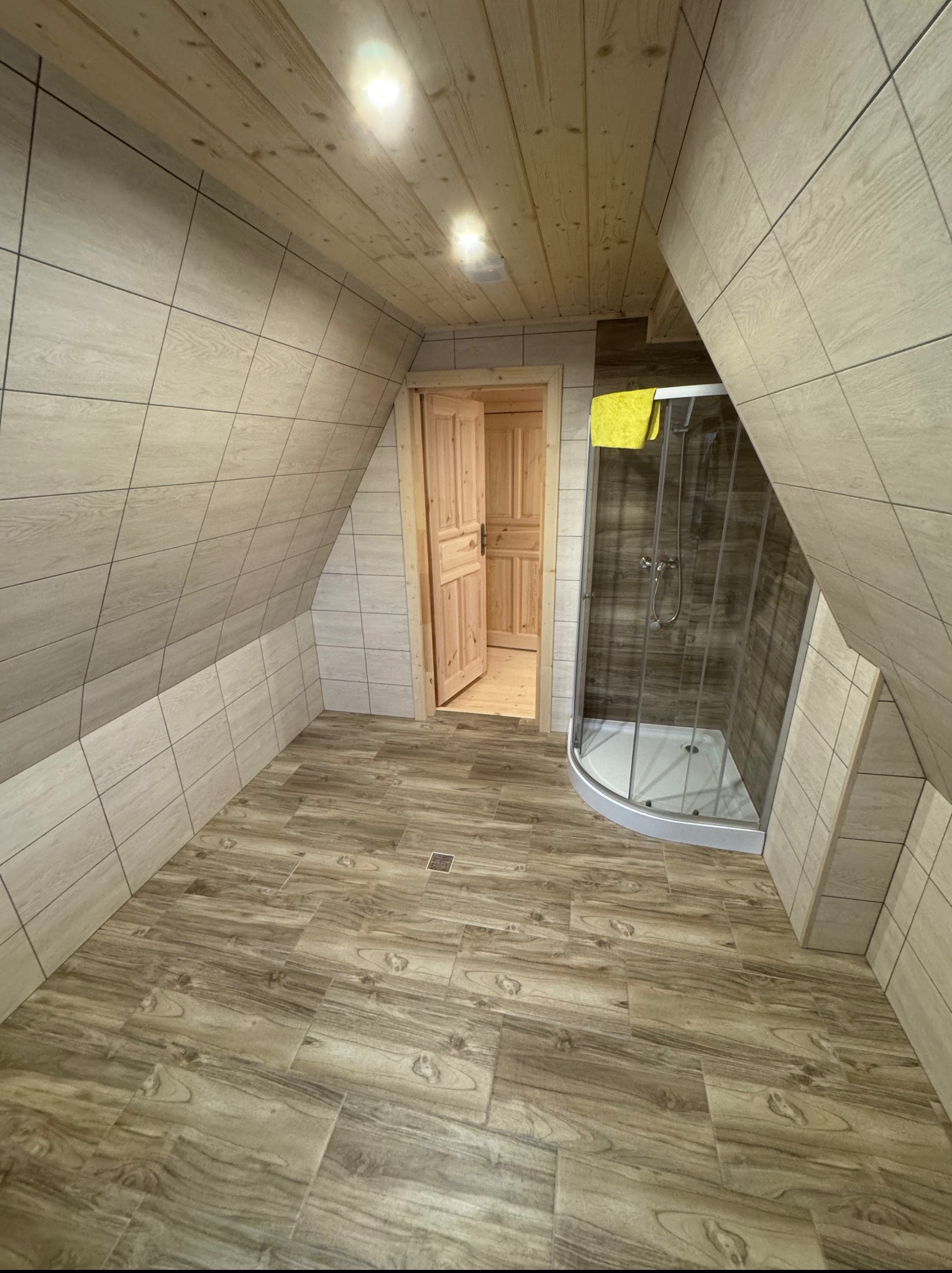 Wykończona łazienka na poddaszu z drewnianą podłogą i kabiną prysznicową. Ściany wyłożone jasnymi płytkami imitującymi drewno. Widoczne drewniane drzwi.