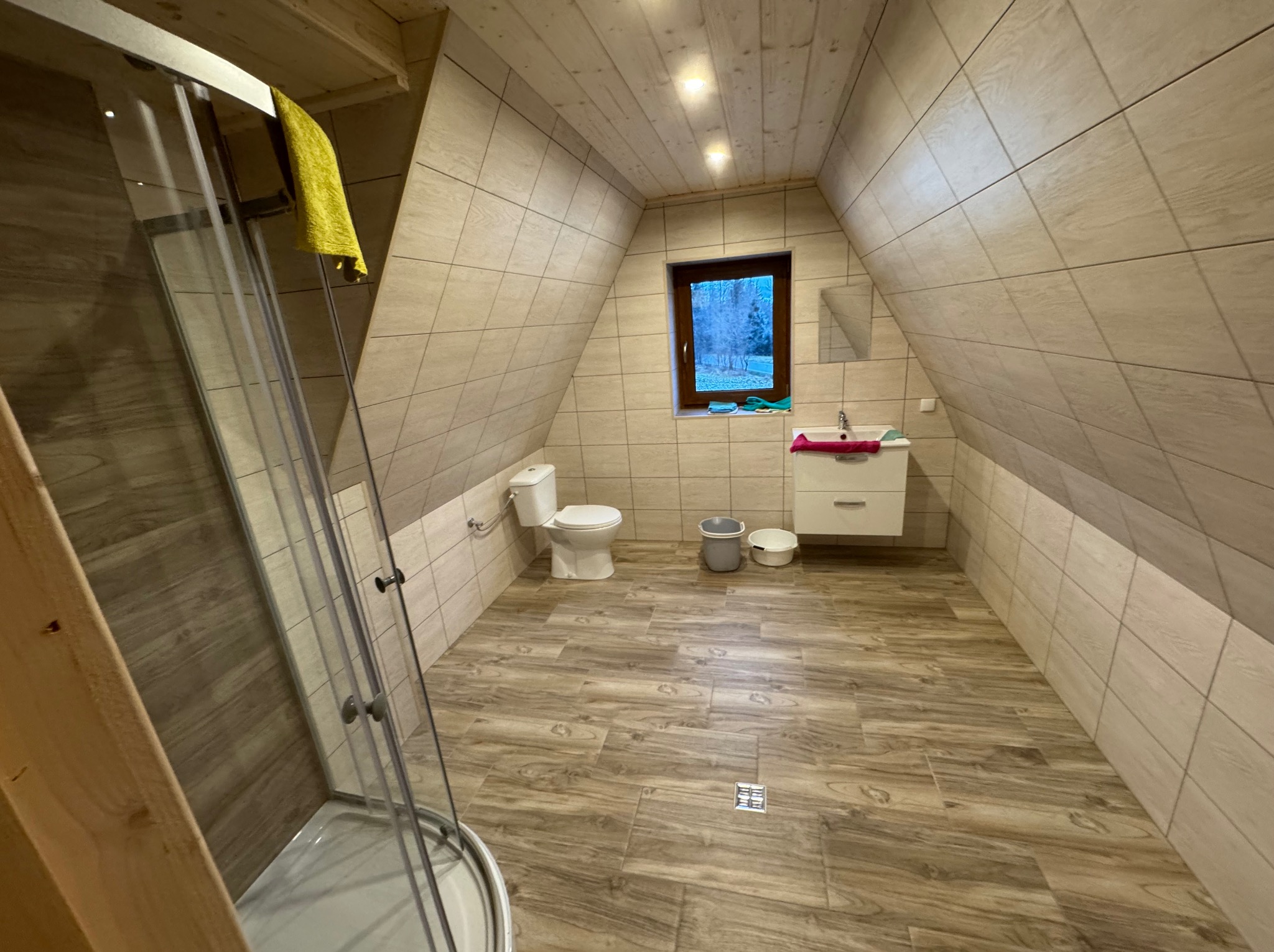 Wykończona łazienka na poddaszu z kabiną prysznicową, toaletą, umywalką z szafką i oknem. Użyto płytek imitujących drewno na podłodze i ścianach.