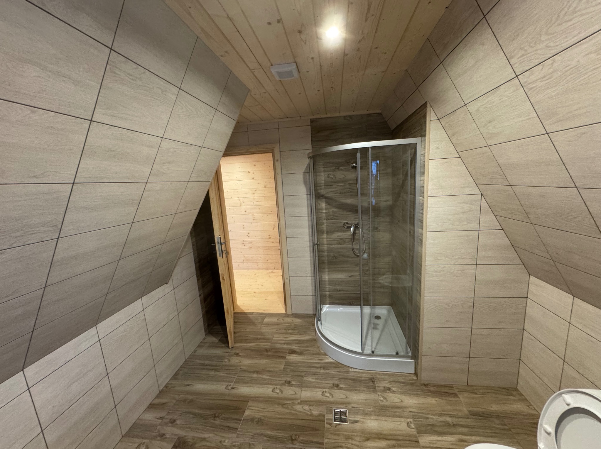 Ukończona łazienka na poddaszu ze skośnymi ścianami wyłożonymi płytkami imitującymi drewno i kabiną prysznicową w rogu. Widoczny fragment toalety.