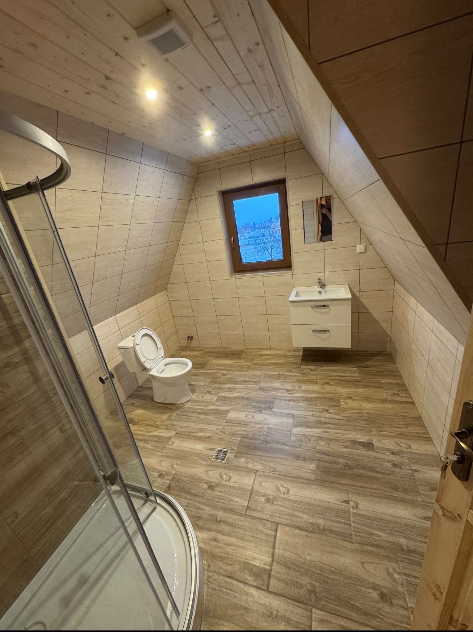 Ukończona łazienka na poddaszu z drewnianym wykończeniem, kabiną prysznicową, toaletą i umywalką z szafką. Okno z widokiem na zimowy krajobraz.
