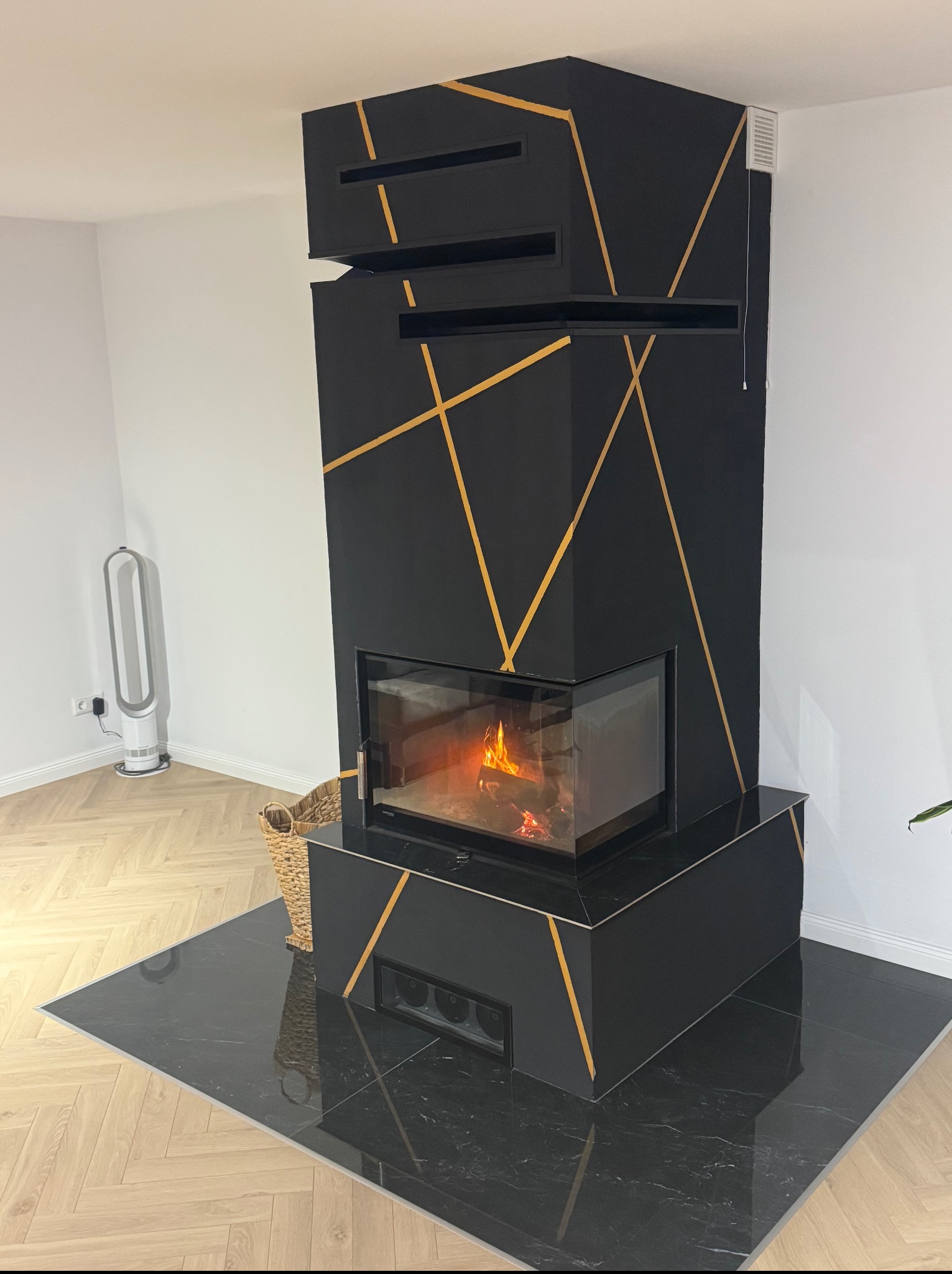 Czarny kominek z geometrycznym, złotym zdobieniem w Ząb. Nowoczesny design, widoczny płomień. Podłoga wyłożona czarnym gresem.
