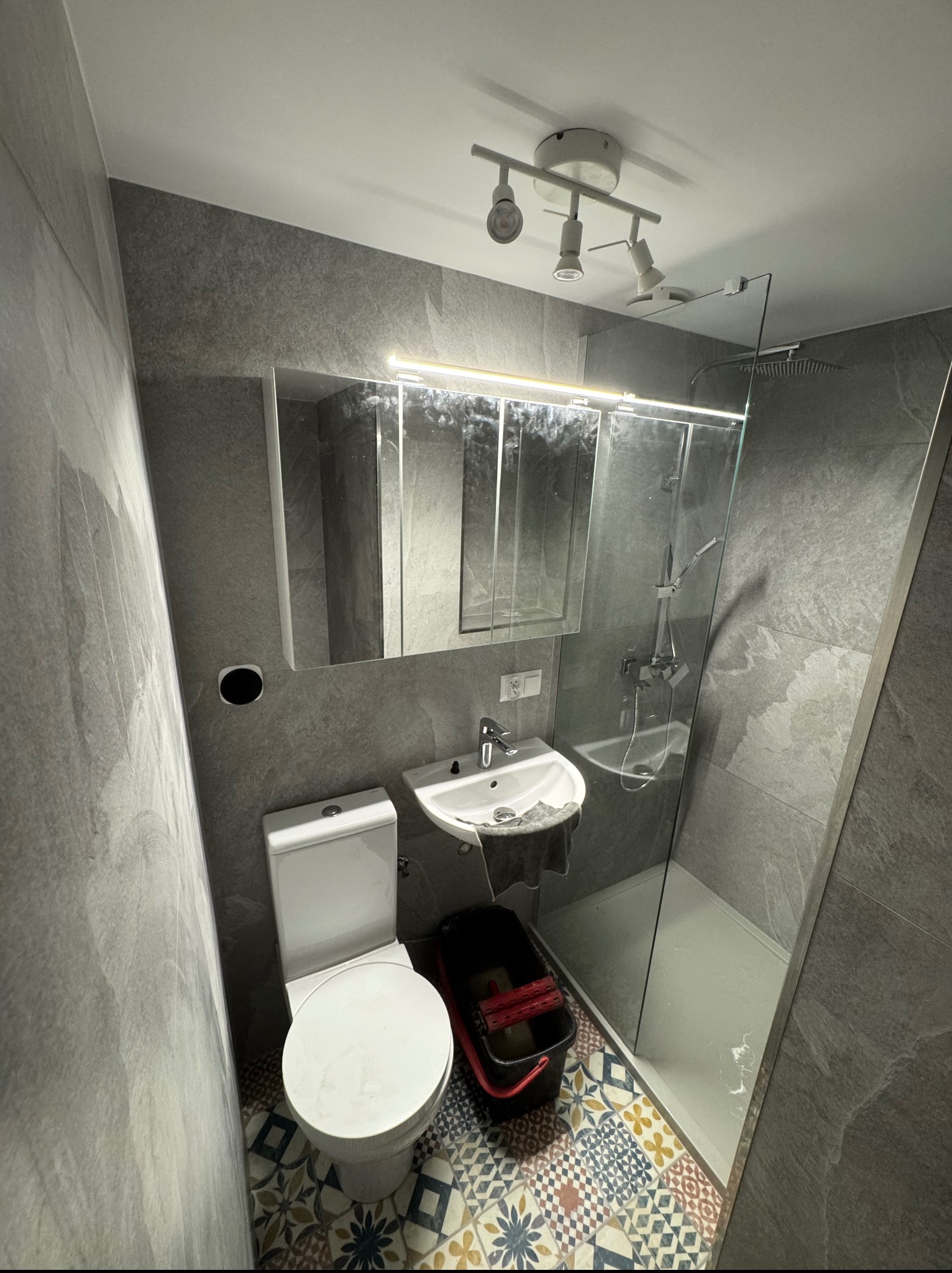 Nowoczesna łazienka po remoncie: szare płytki imitujące kamień na ścianach, kabina prysznicowa ze szkła, toaleta, umywalka i kolorowa mozaika na podłodze.