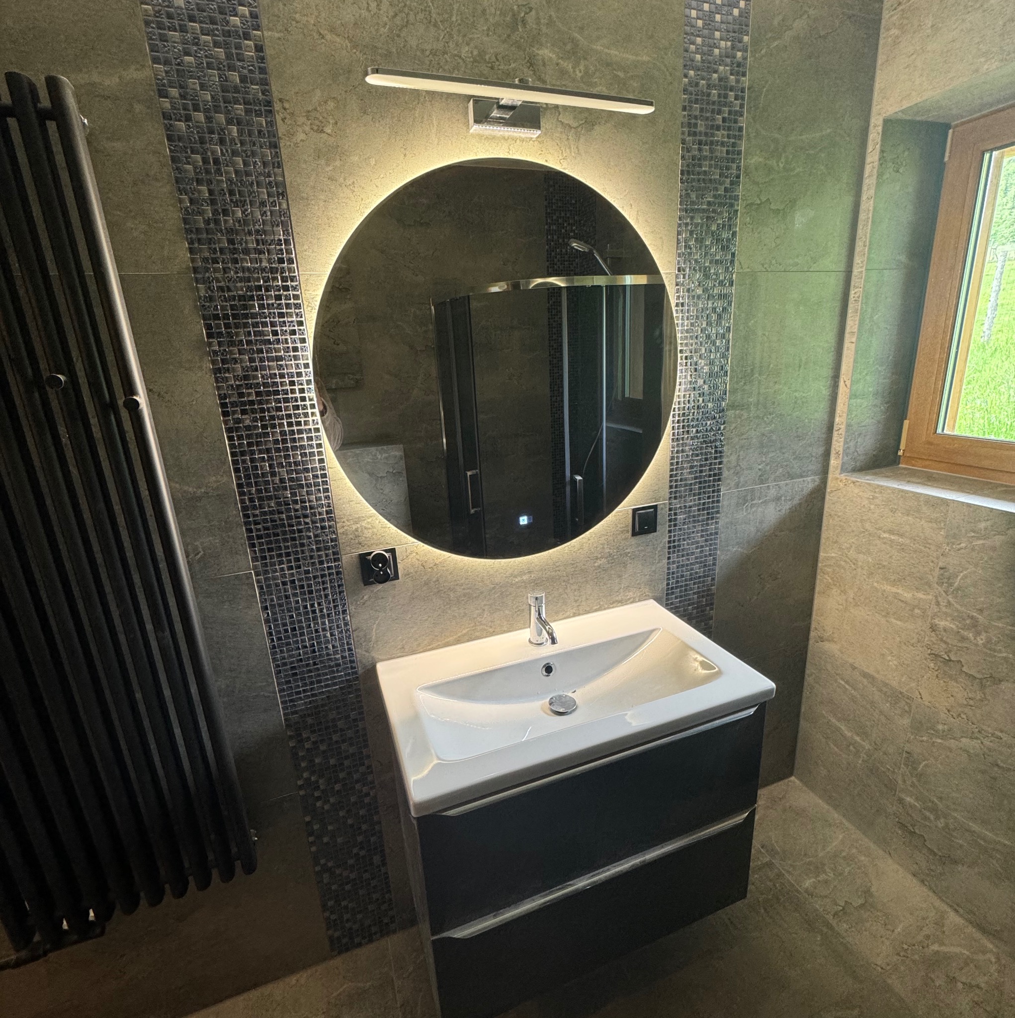 Nowoczesna łazienka z umywalką nablatową, okrągłym lustrem z podświetleniem LED i grzejnikiem drabinkowym. Wykończenie w odcieniach szarości z mozaiką.