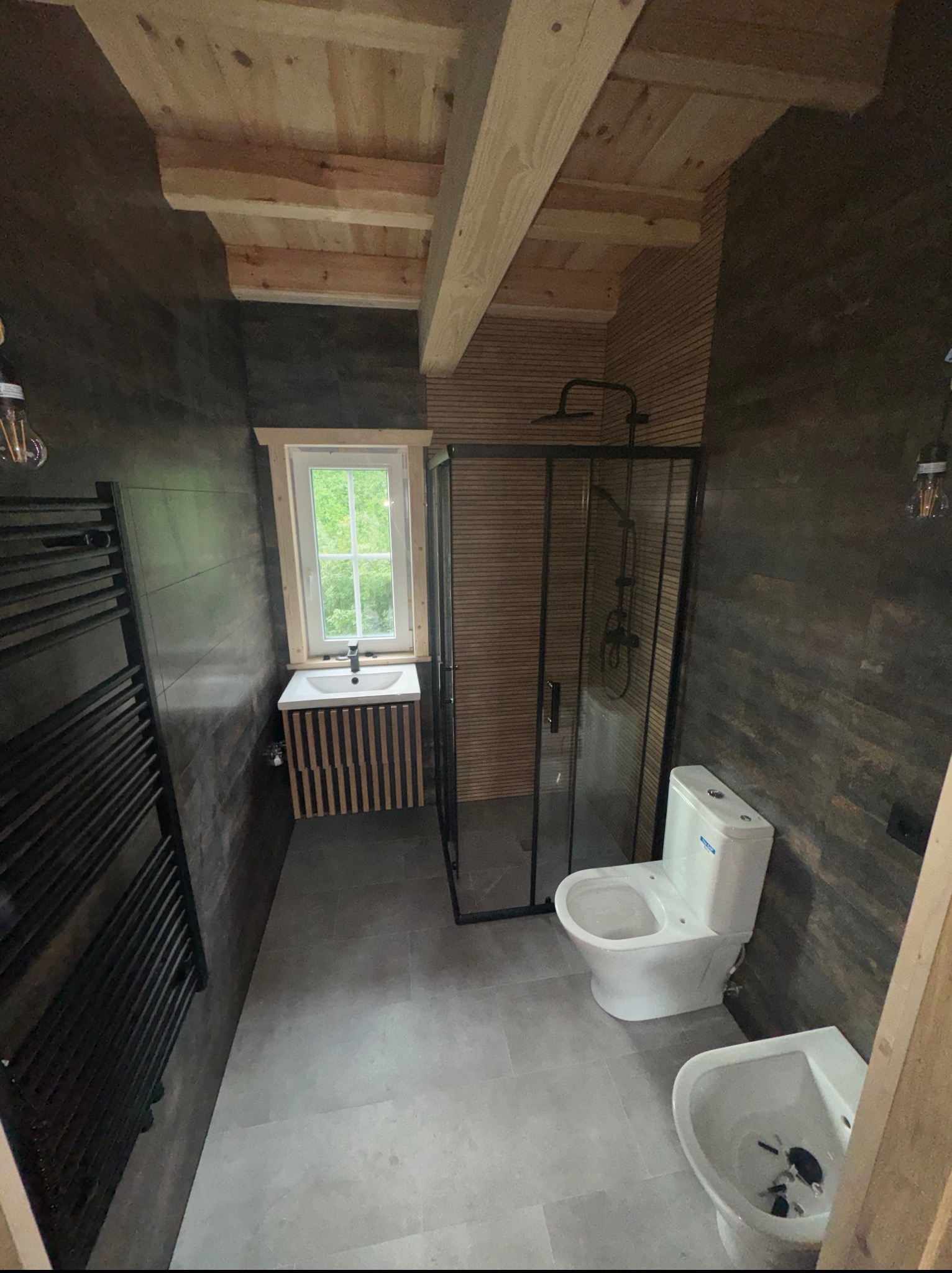 Nowoczesna łazienka z kabiną prysznicową typu walk-in, umywalką z szafką z drewnianymi lamelami i grzejnikiem drabinkowym na tle ciemnych, drewnopodobnych płytek.