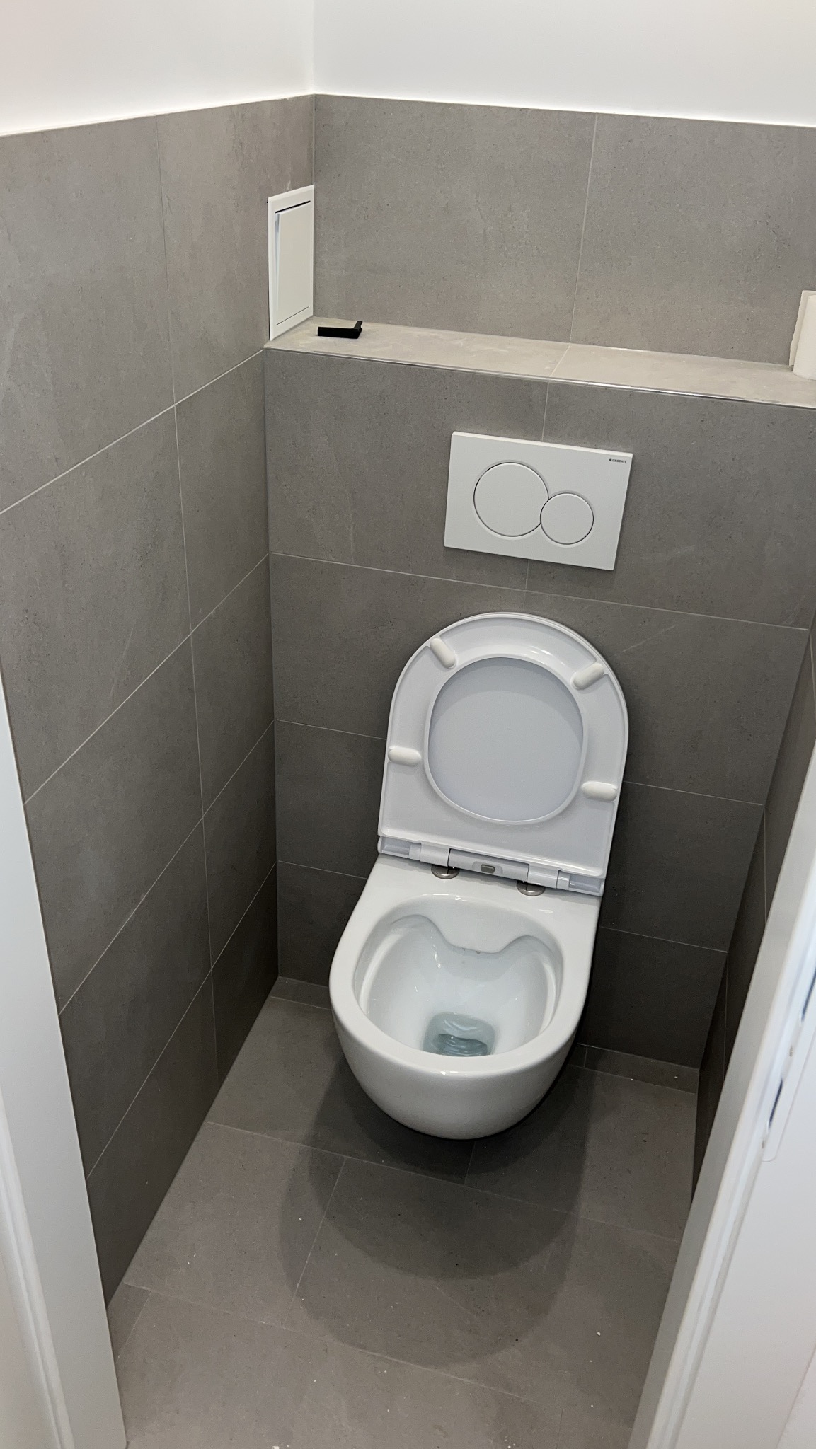 Nowoczesna toaleta w szarej łazience z płytkami. Podtynkowa spłuczka Geberit i dostęp rewizyjny. Minimalistyczny design.