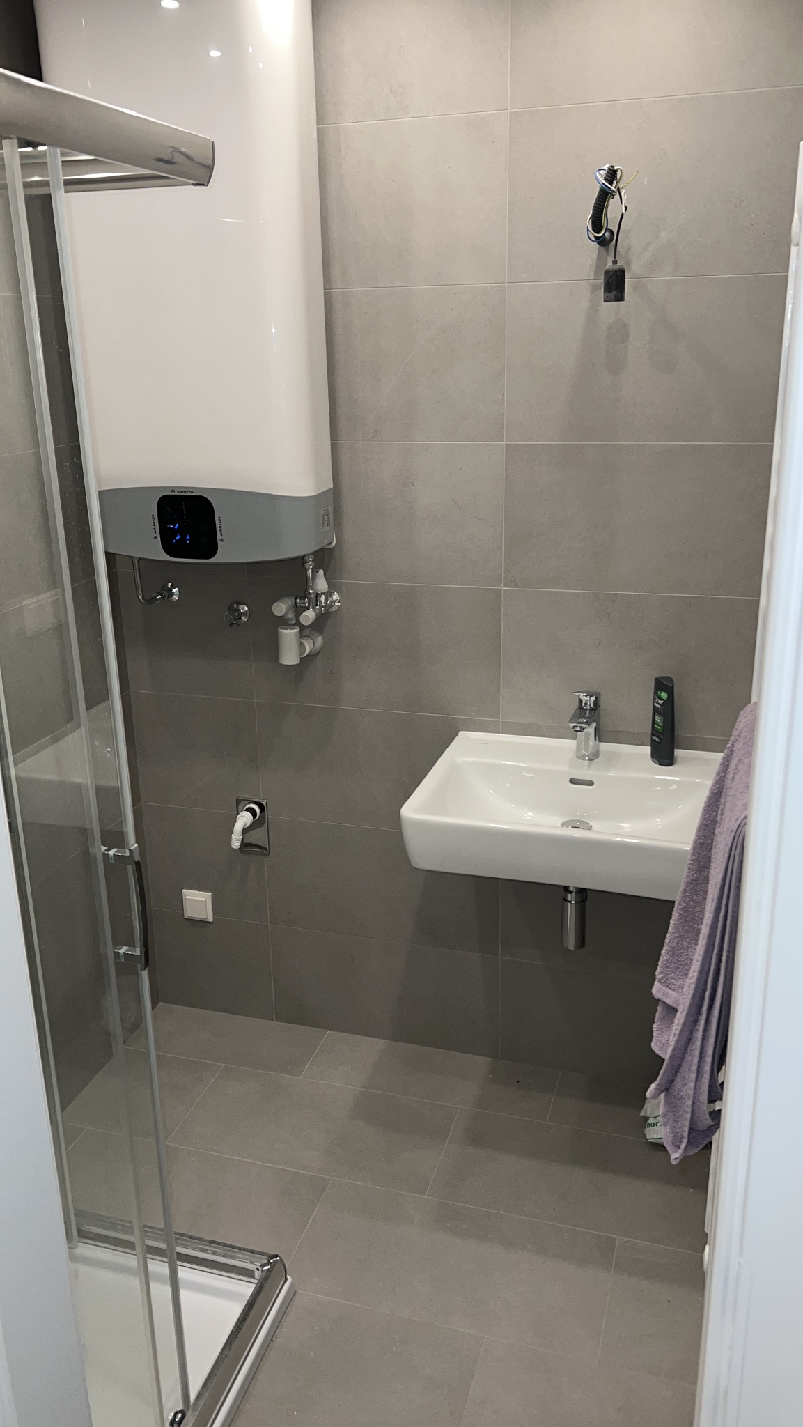 Nowoczesna łazienka z szarymi płytkami, kabiną prysznicową, bojlerem i umywalką. Minimalistyczny design, widoczne przyłącza i armatura.