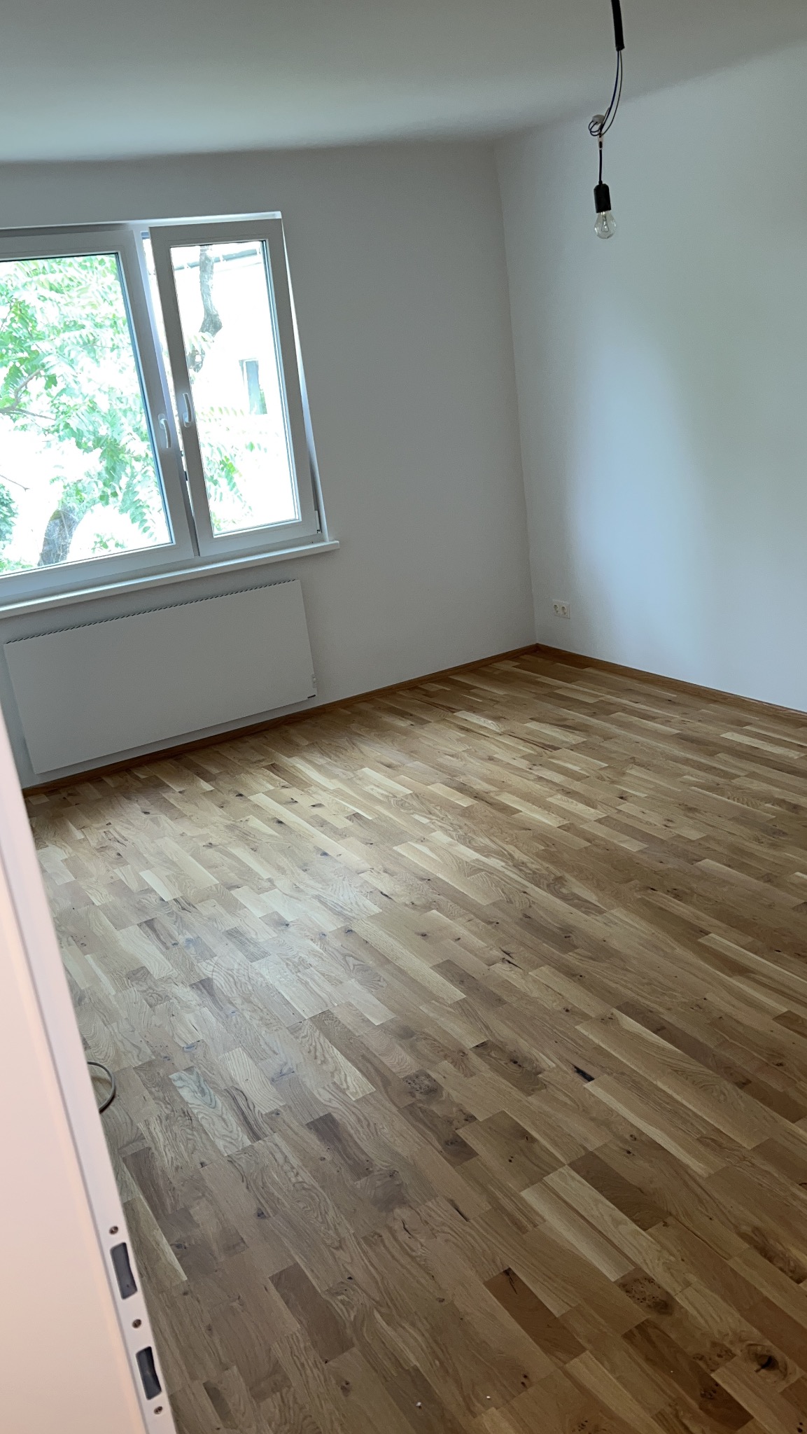 Jasne, drewniane panele podłogowe w pokoju z białymi ścianami i grzejnikiem pod oknem. Wisząca żarówka dodaje surowego charakteru wnętrzu.