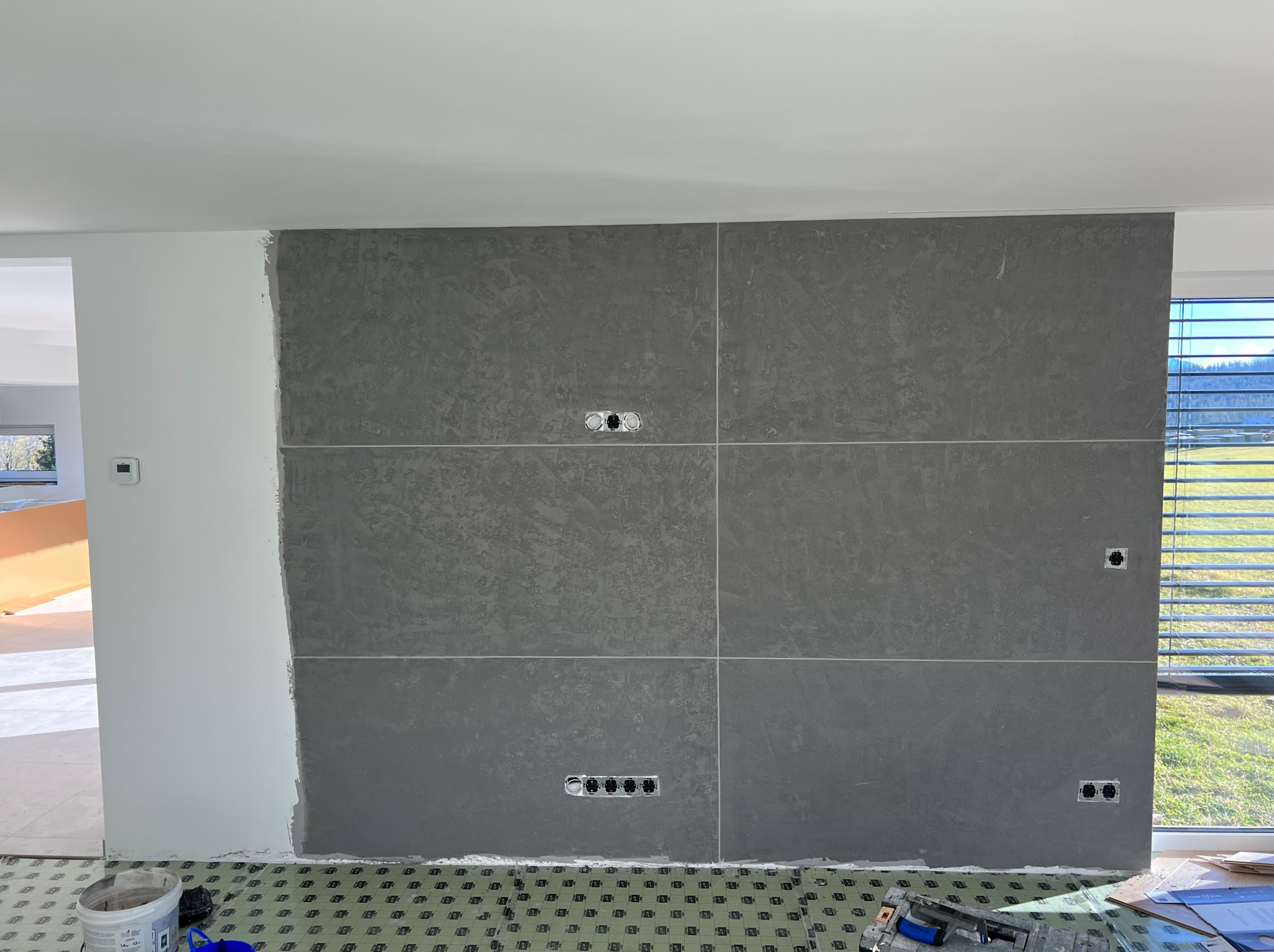 Wykończona ściana z szarych płyt imitujących kamień, z widocznymi gniazdkami elektrycznymi. Na podłodze folia ochronna, a obok narzędzia glazurnicze.