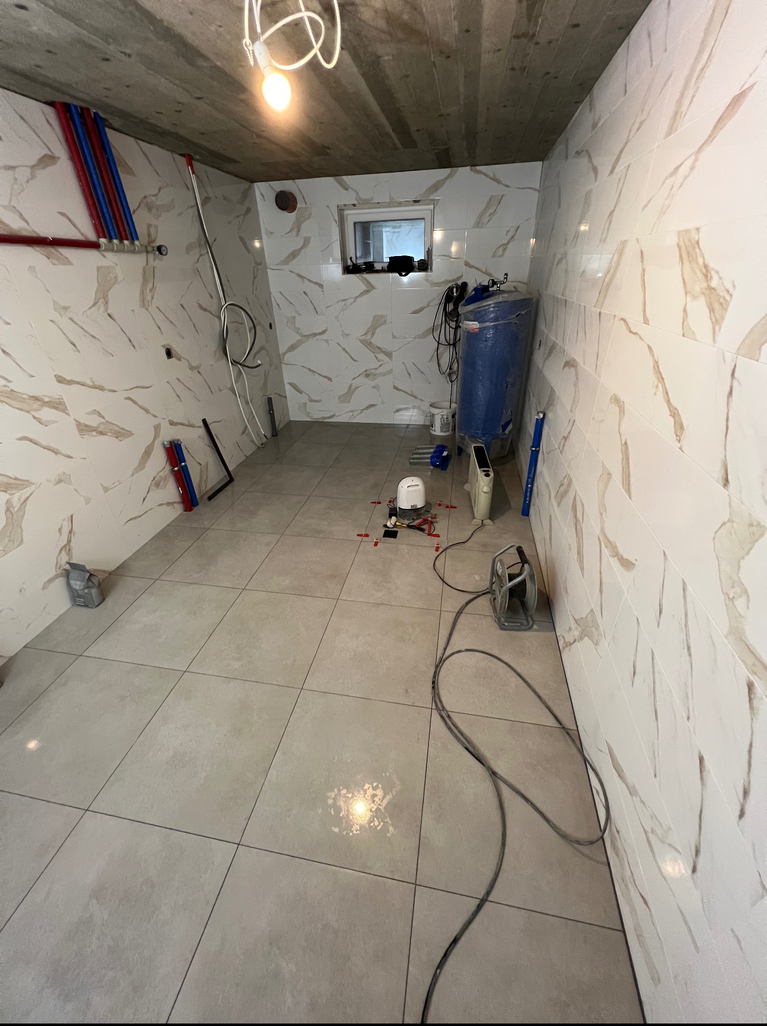 Wnętrze łazienki w trakcie prac wykończeniowych: ułożone płytki podłogowe i ścienne imitujące marmur, widoczne rury, bojler i narzędzia glazurnicze pod żarówką.