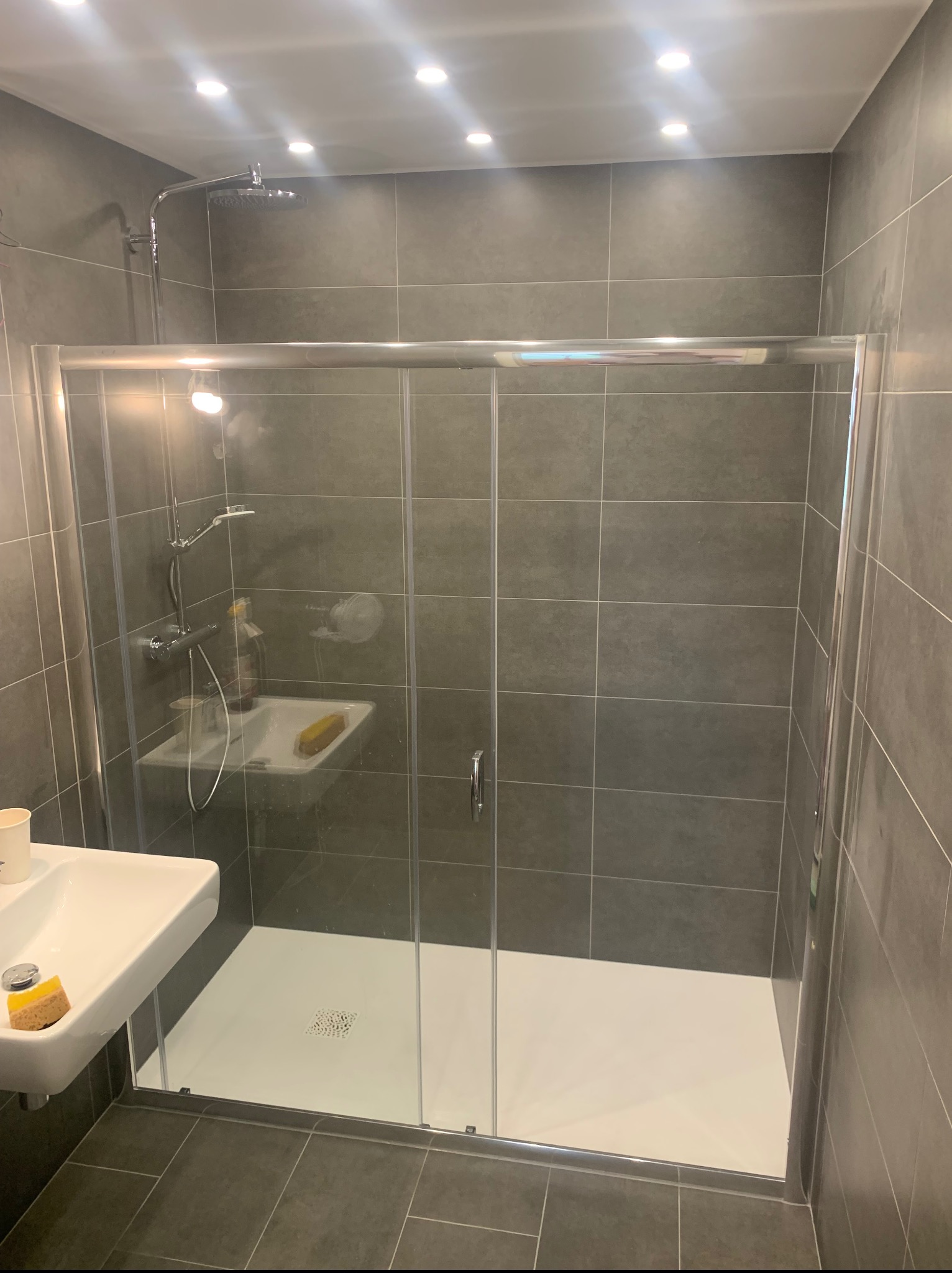 Nowoczesna kabina prysznicowa z przesuwnymi drzwiami, wykończona szarą płytką, z widoczną armaturą i umywalką. Minimalistyczny design, oświetlenie punktowe.