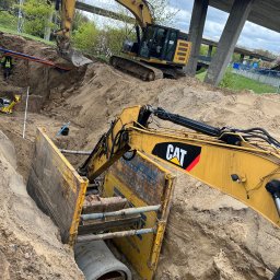 AQUA WIK SPÓŁKA Z OGRANICZONĄ ODPOWIEDZIALNOŚCIĄ - Żółty koparka CAT w wykopie zabezpieczonym szalunkiem, w tle druga koparka i konstrukcja mostu. Widoczne rury i pracownik w odblaskowej kamizelce.