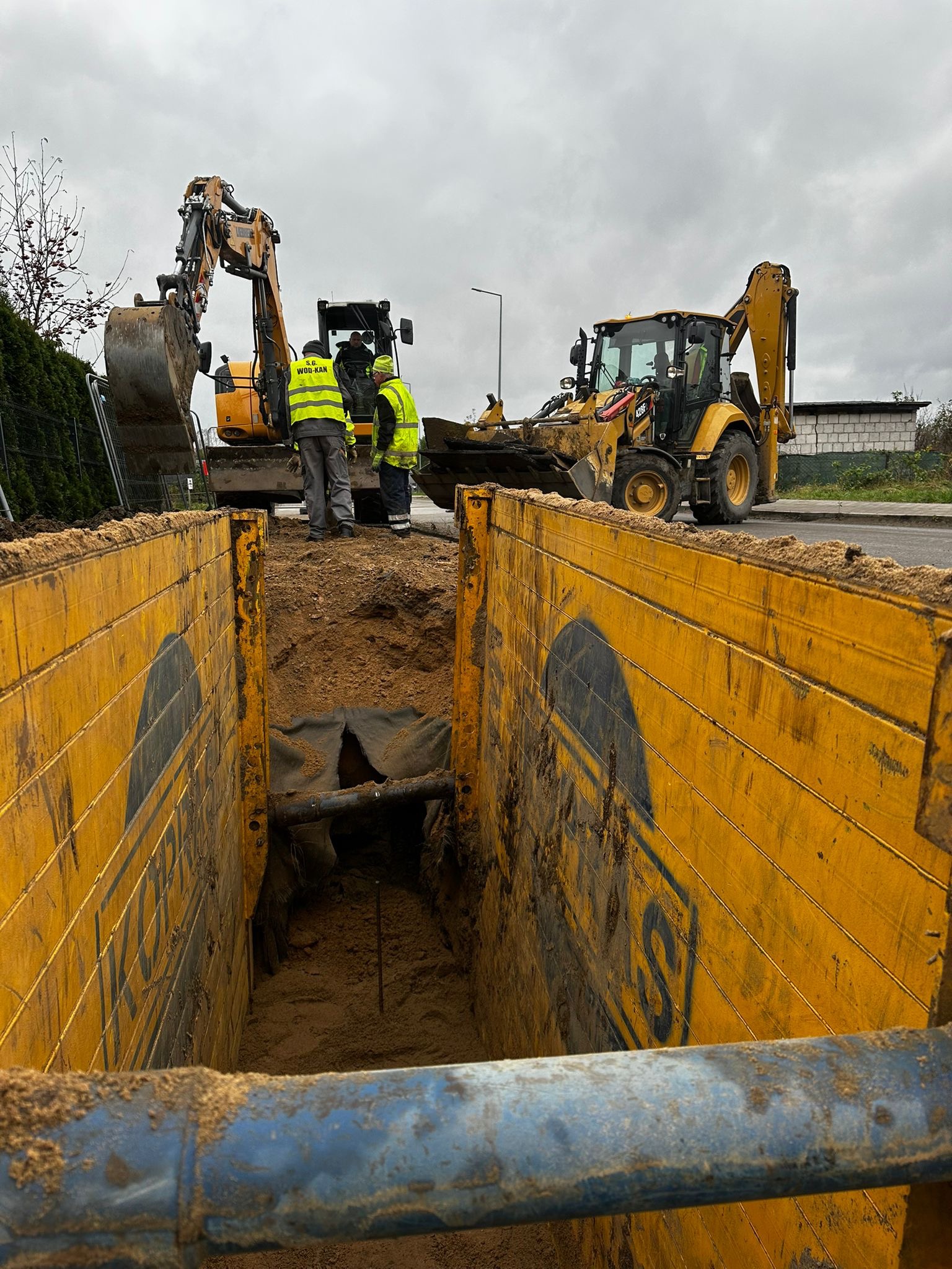 Głęboki wykop zabezpieczony żółtymi szalunkami, z widocznymi koparkami i pracownikami w odblaskowych kamizelkach. Wykop przygotowany pod instalacje, w tle budynek.