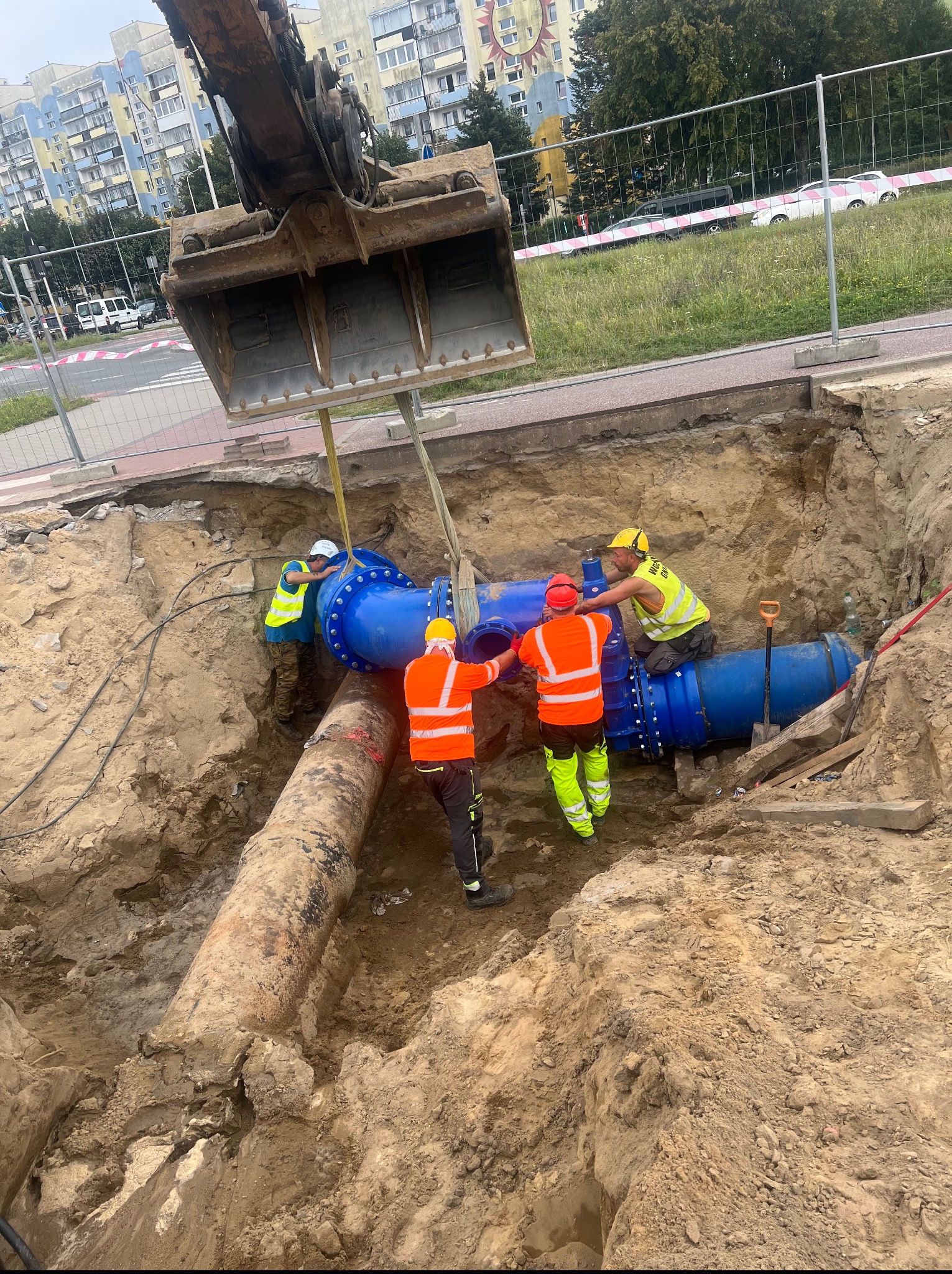 Montaż nowego, niebieskiego rurociągu w wykopie przez grupę robotników w kaskach i kamizelkach odblaskowych przy użyciu koparki na tle miejskiego krajobrazu.