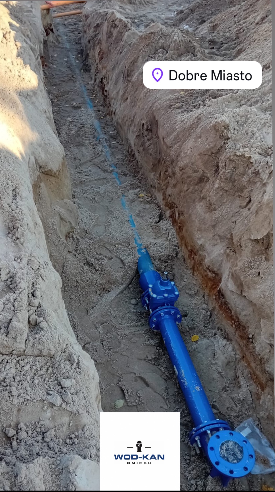 Niebieska rura wodociągowa z zaworem, ułożona w wykopie w ziemi. Widoczny fragment rury na całej długości wykopu. Logo firmy WOD-KAN Gniech.