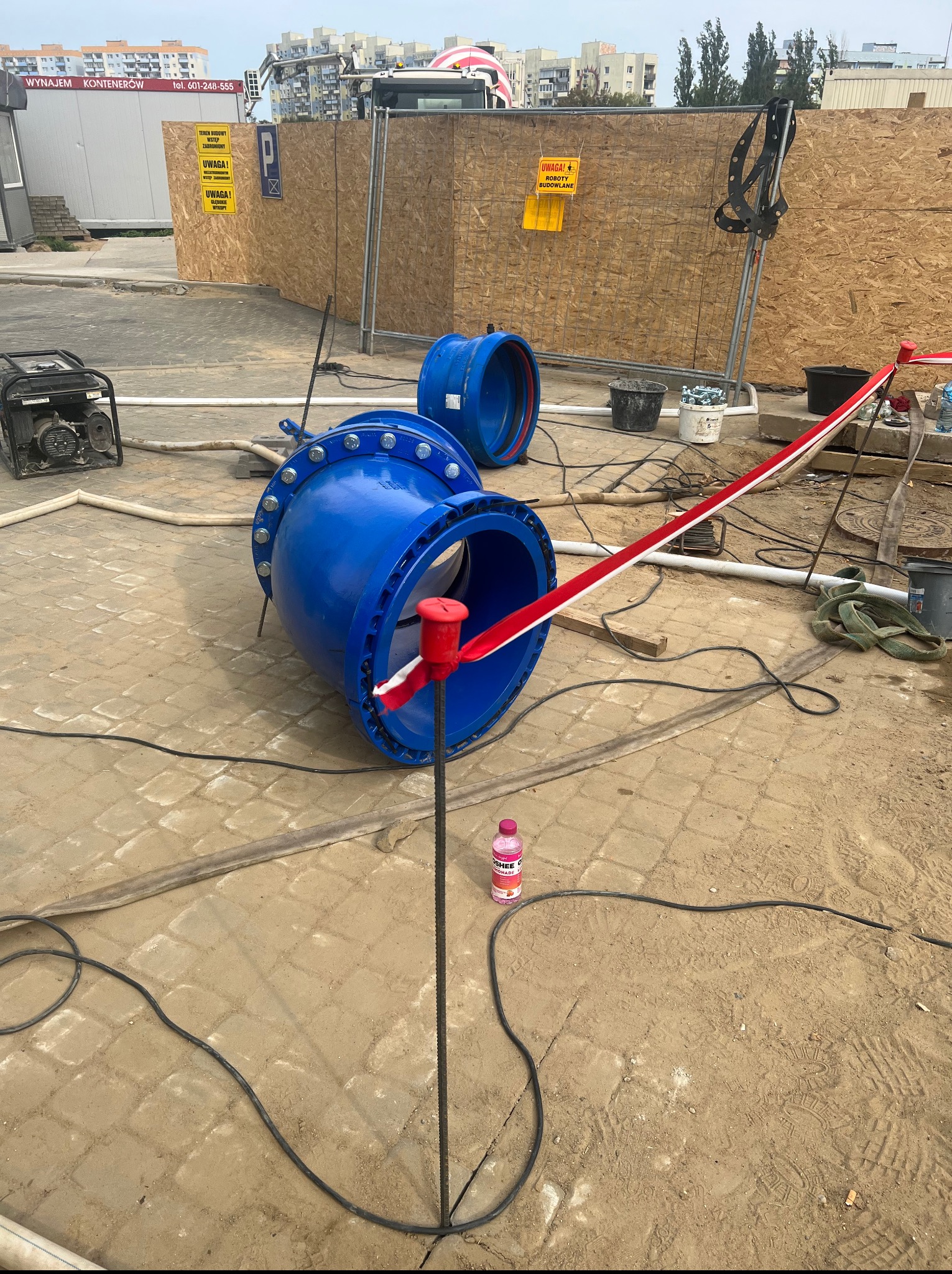 Niebieskie rury z kołnierzami na placu budowy, otoczone taśmą ostrzegawczą i narzędziami. Widoczne ogrodzenie budowlane i budynki w tle.