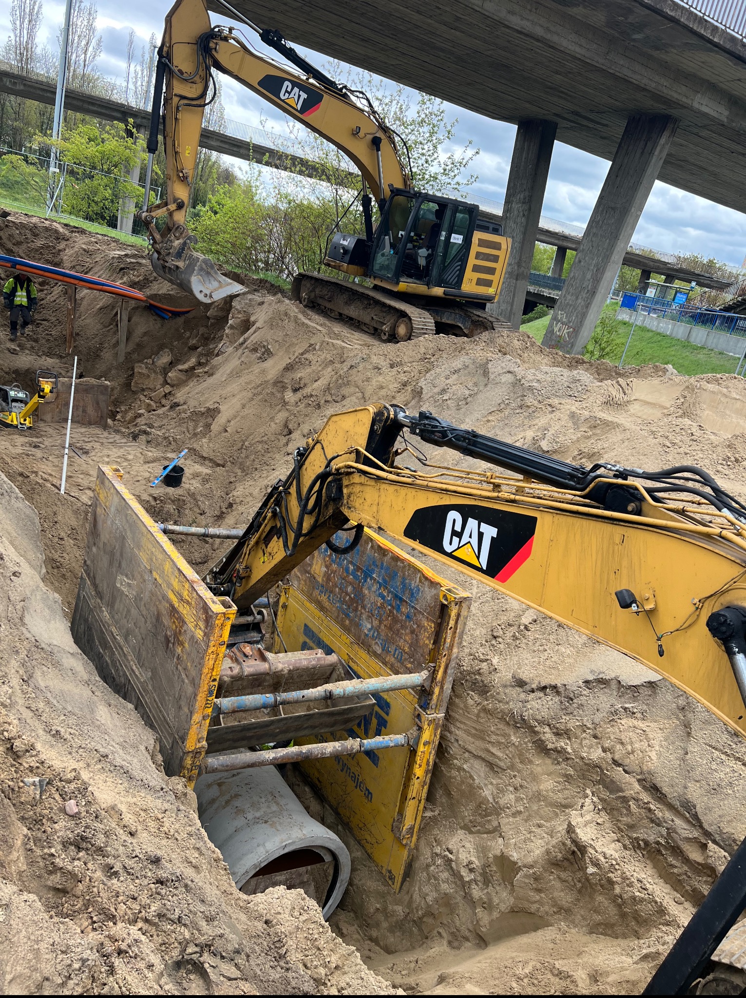 Żółty koparka CAT w wykopie zabezpieczonym szalunkiem, w tle druga koparka i konstrukcja mostu. Widoczne rury i pracownik w odblaskowej kamizelce.