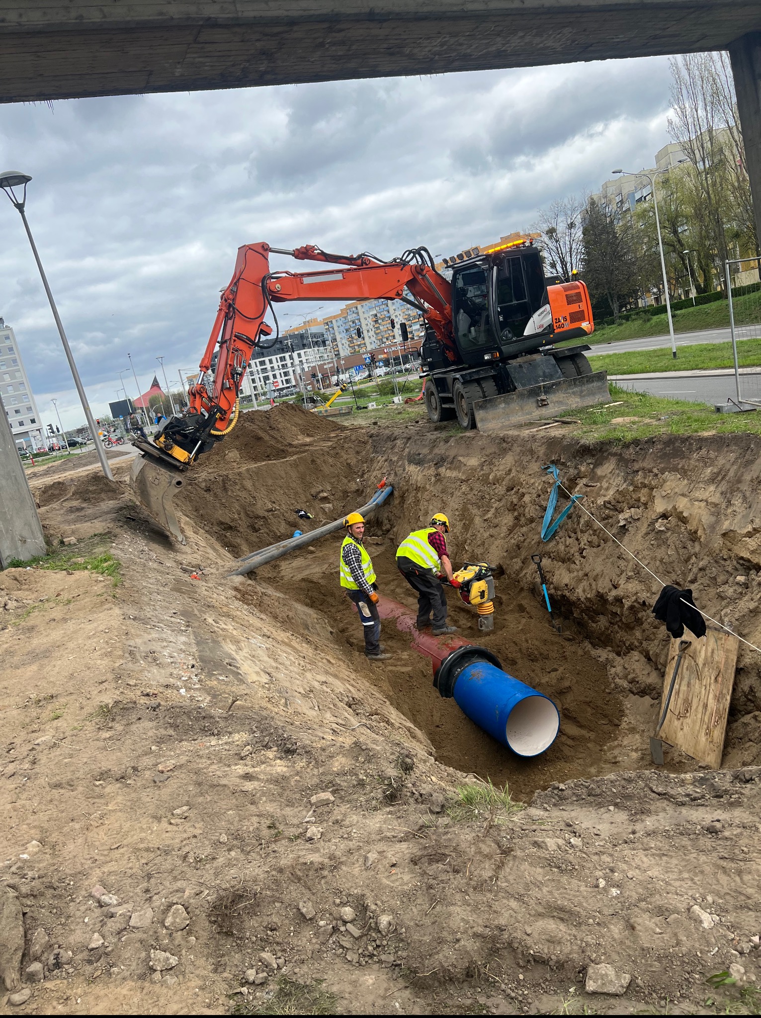 Wykonawca w wykopie zagęszcza podłoże pod rury, w tle pomarańczowa koparka. Prace ziemne i instalacyjne na tle miejskiego krajobrazu.