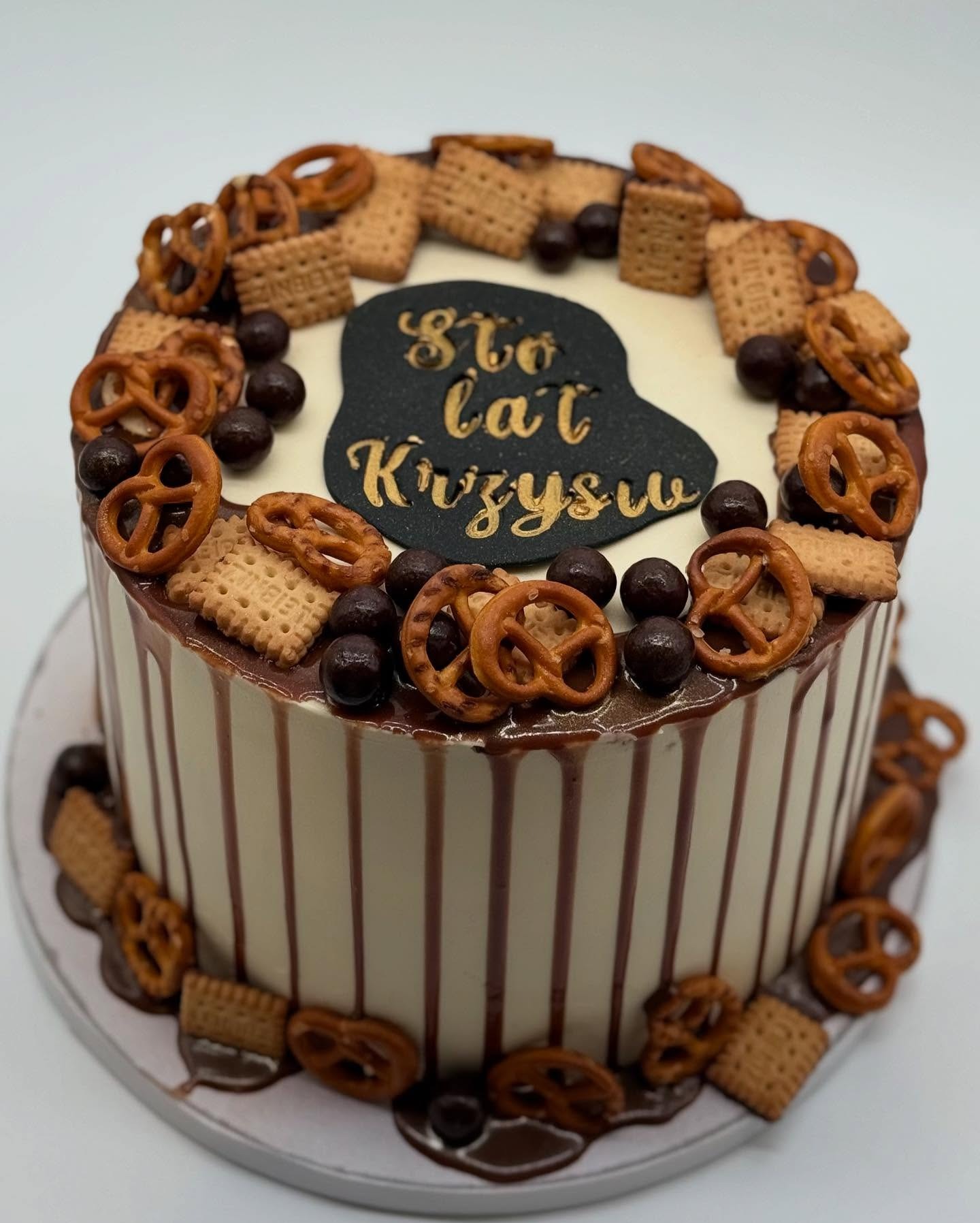 Tort urodzinowy z napisem 'Sto lat Krzysiu', dekorowany preclami, ciastkami i czekoladowymi kulkami. Kremowe tło i brązowe polewy.