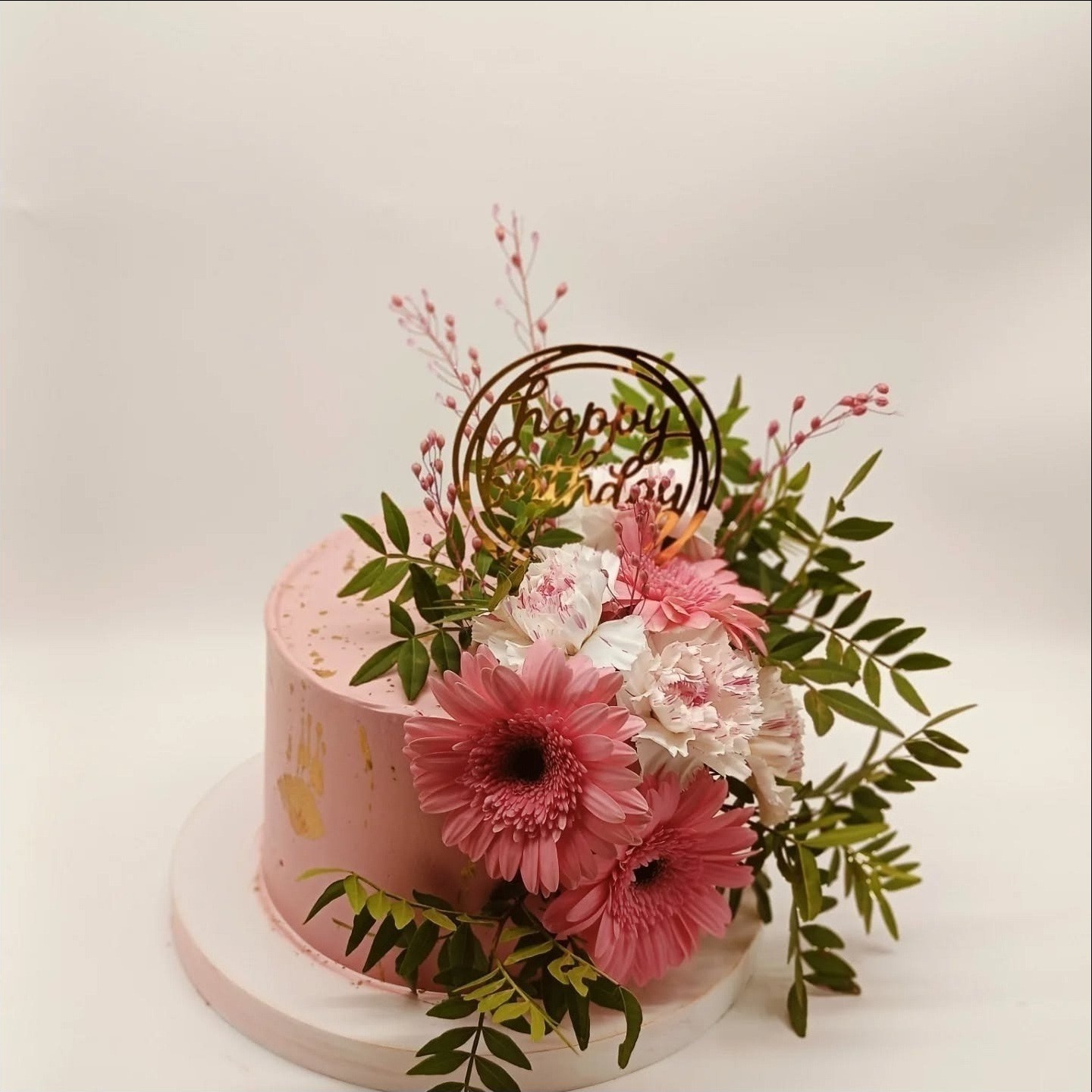 Tort urodzinowy w odcieniach różu, zdobiony świeżymi kwiatami i złotym topperem 'Happy Birthday', na białym tle. Delikatne zdobienia złotem na torcie.