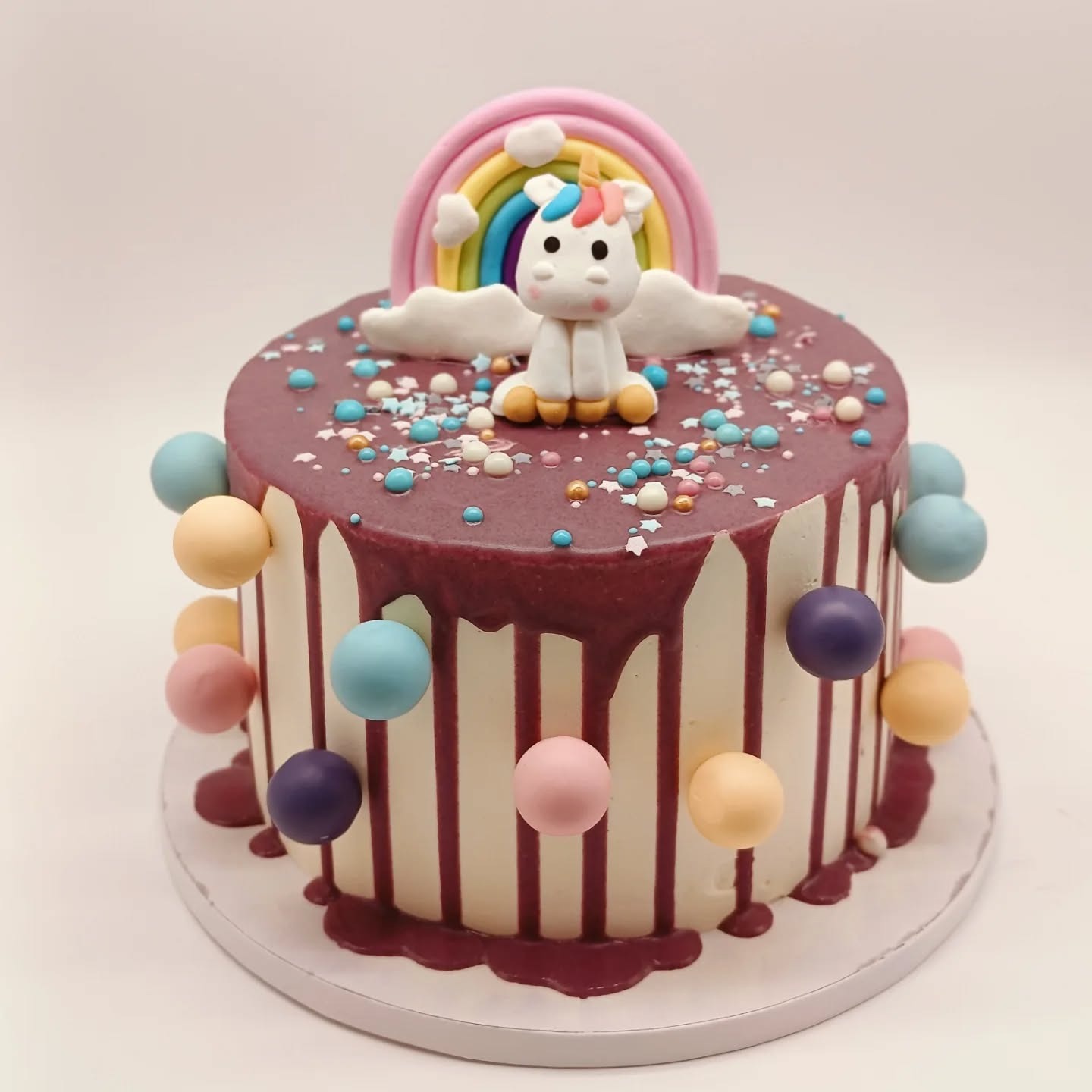 Okrągły tort z polewą, dekorowany tęczą, jednorożcem i kolorowymi kuleczkami. Kremowe tło i starannie wykonane detale.