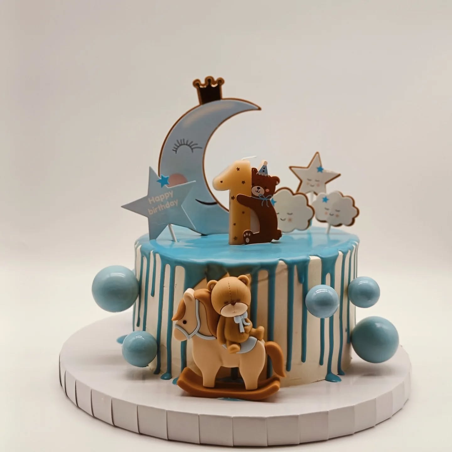 Udekorowany tort urodzinowy dla roczniaka z motywami księżyca, gwiazd, chmurek i misia na koniku, z niebieskim lukrem i dekoracjami.
