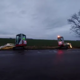 Dwa mini-koparki na skraju pola, jedna z włączonym światłem, wykonujące wykop w pochmurny dzień. W tle turbiny wiatrowe i samotne drzewo.