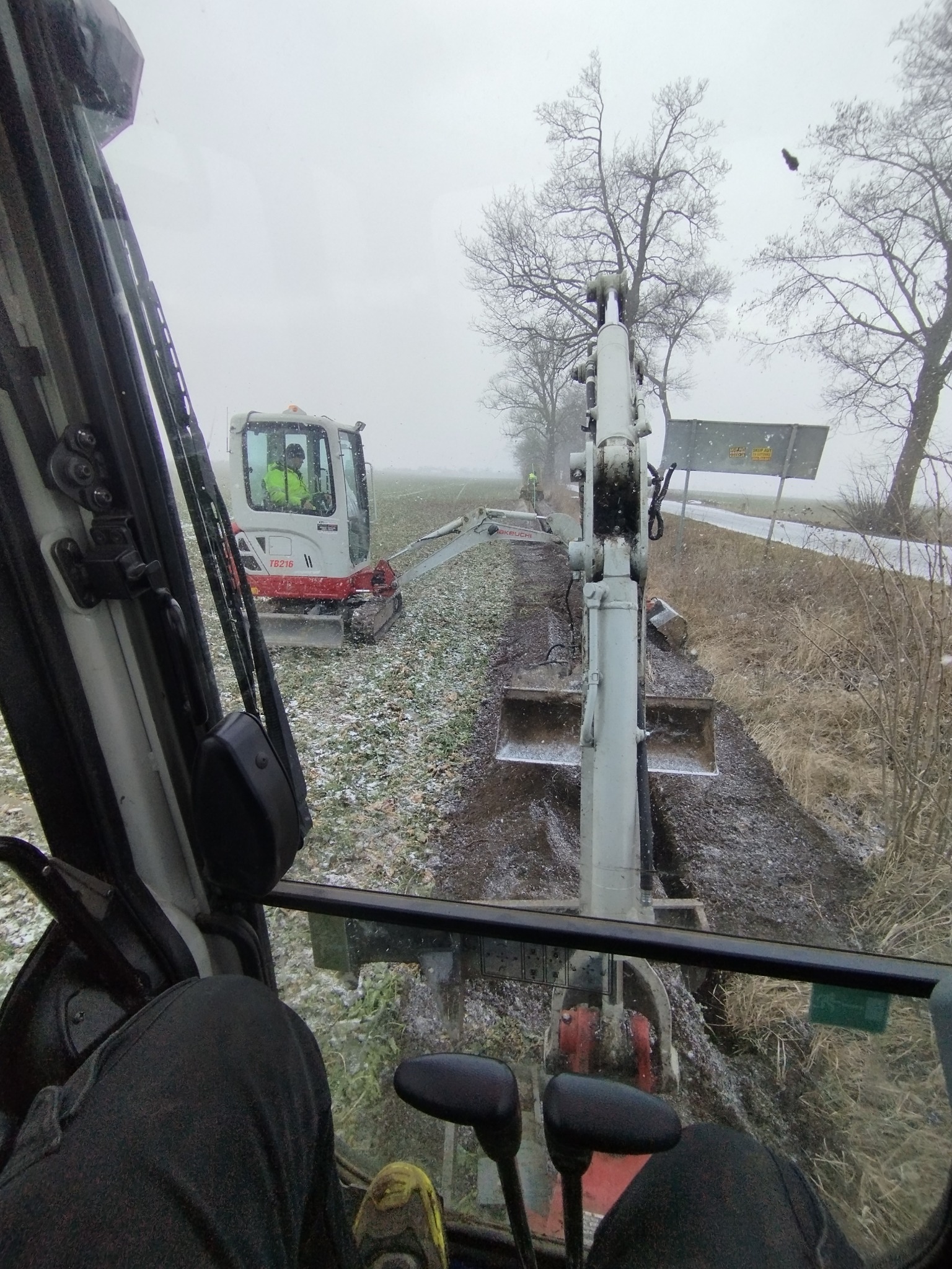 Widok z kabiny koparki na prace ziemne w terenie pokrytym śniegiem. W tle widoczna druga koparka i operator w odblaskowej kamizelce. Pochmurne niebo.