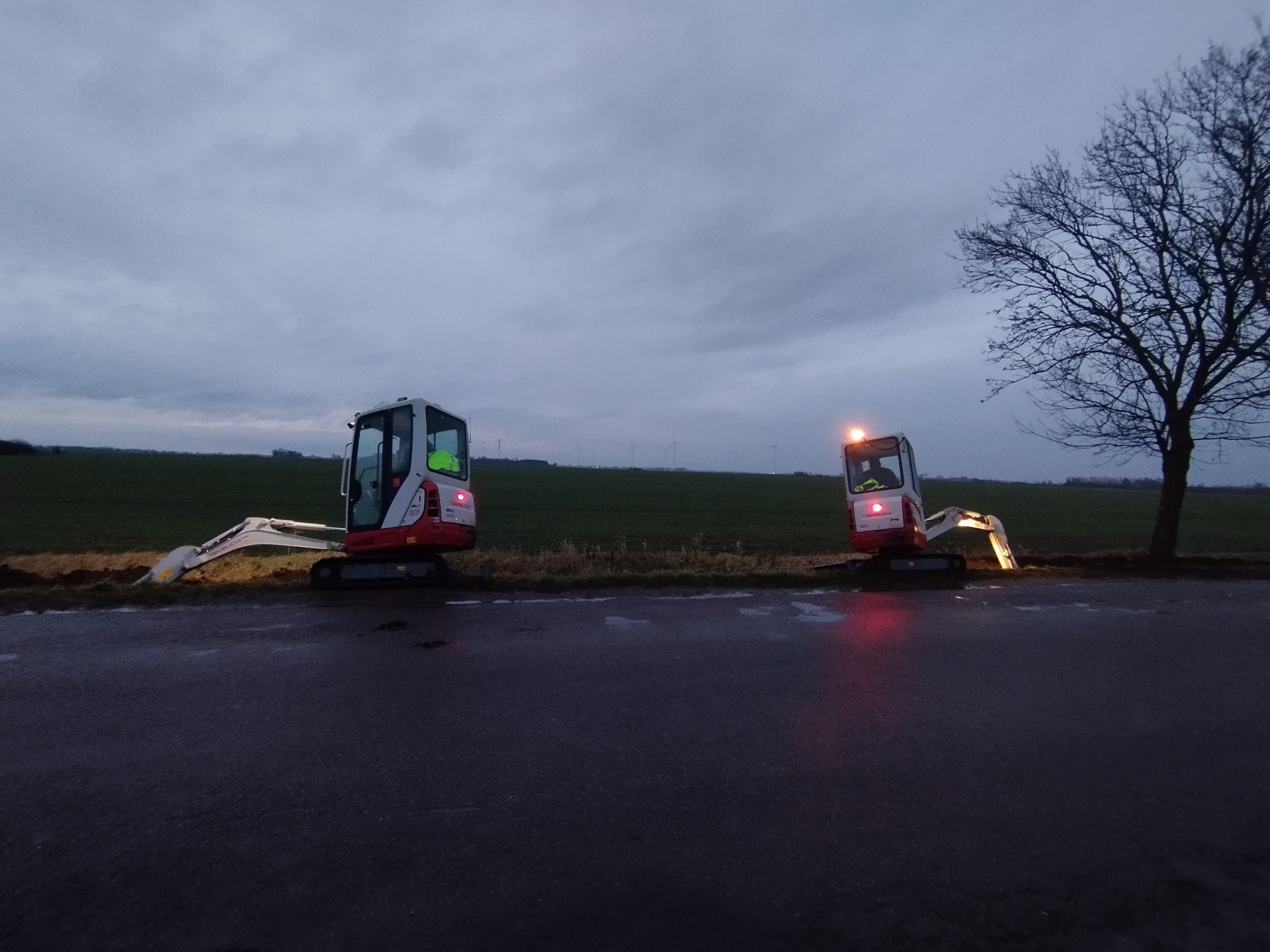 Dwa mini-koparki na skraju pola, jedna z włączonym światłem, wykonujące wykop w pochmurny dzień. W tle turbiny wiatrowe i samotne drzewo.
