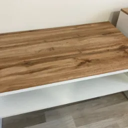 Zmontowany stolik kawowy z blatem imitującym drewno i białymi półkami, ustawiony na panelach podłogowych. Widoczny fragment drugiego mebla w tle.