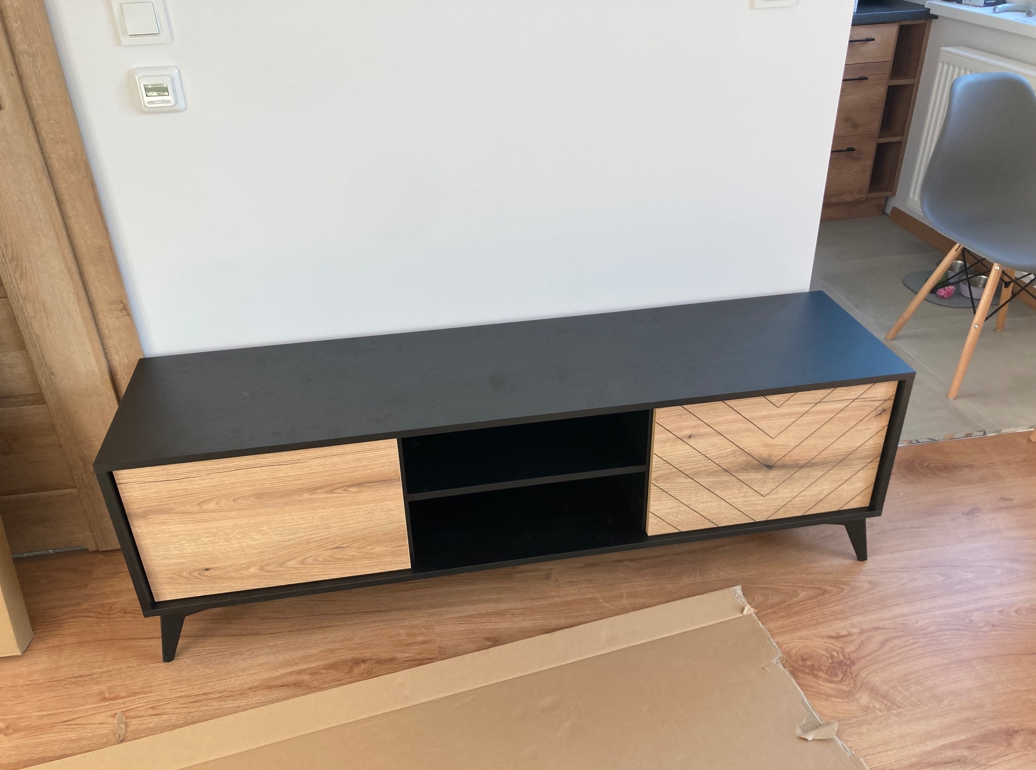 Czarno-drewniana szafka RTV z wzorem jodełki na frontach, ustawiona na panelach. W tle widoczny fragment ściany z włącznikiem i regulator temperatury.