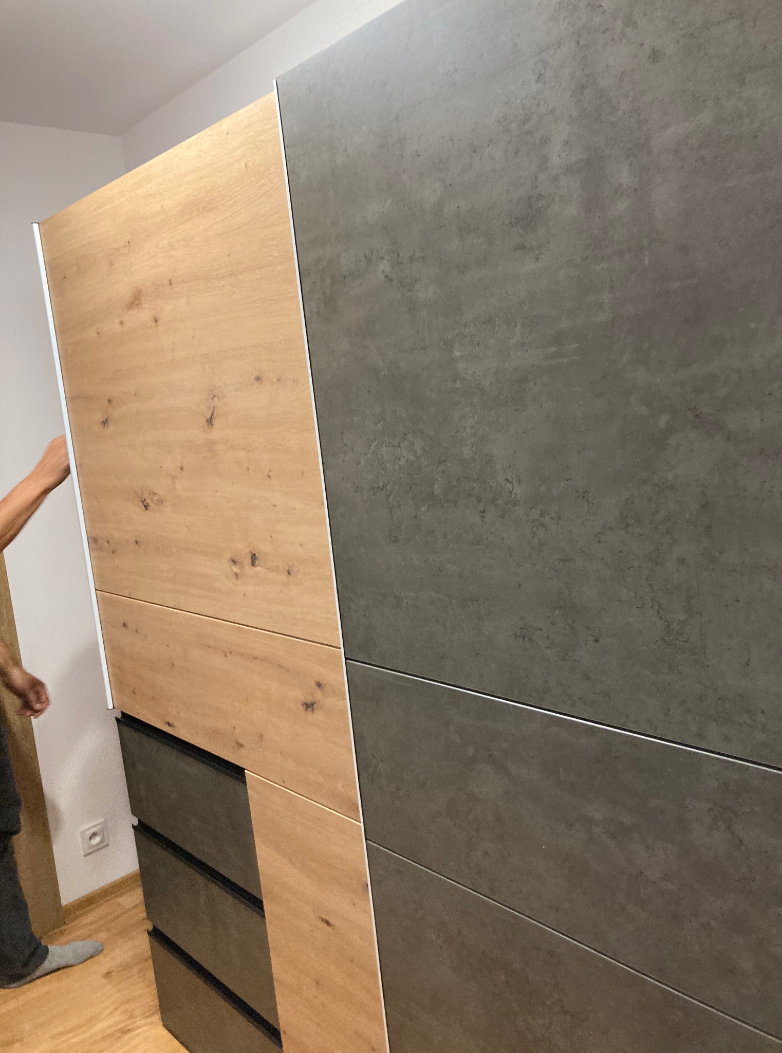 Nowoczesna szafa przesuwna z drewnianym i szarym panelem, widoczna część sylwetki osoby podczas montażu, wnętrze pomieszczenia.