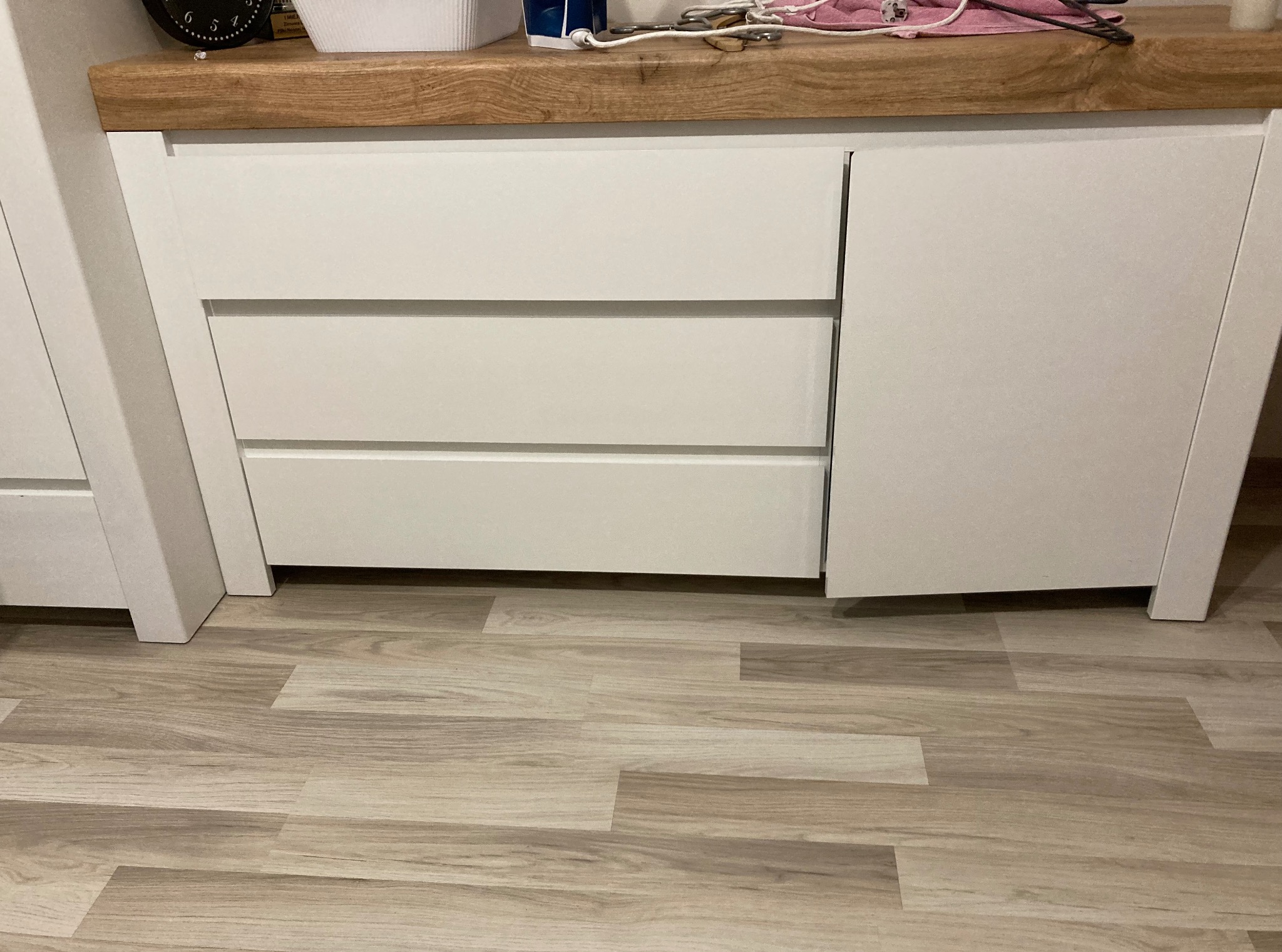 Biała szafka z trzema szufladami i drewnianym blatem, ustawiona na panelach podłogowych. Drzwiczki szafki są lekko uchylone.