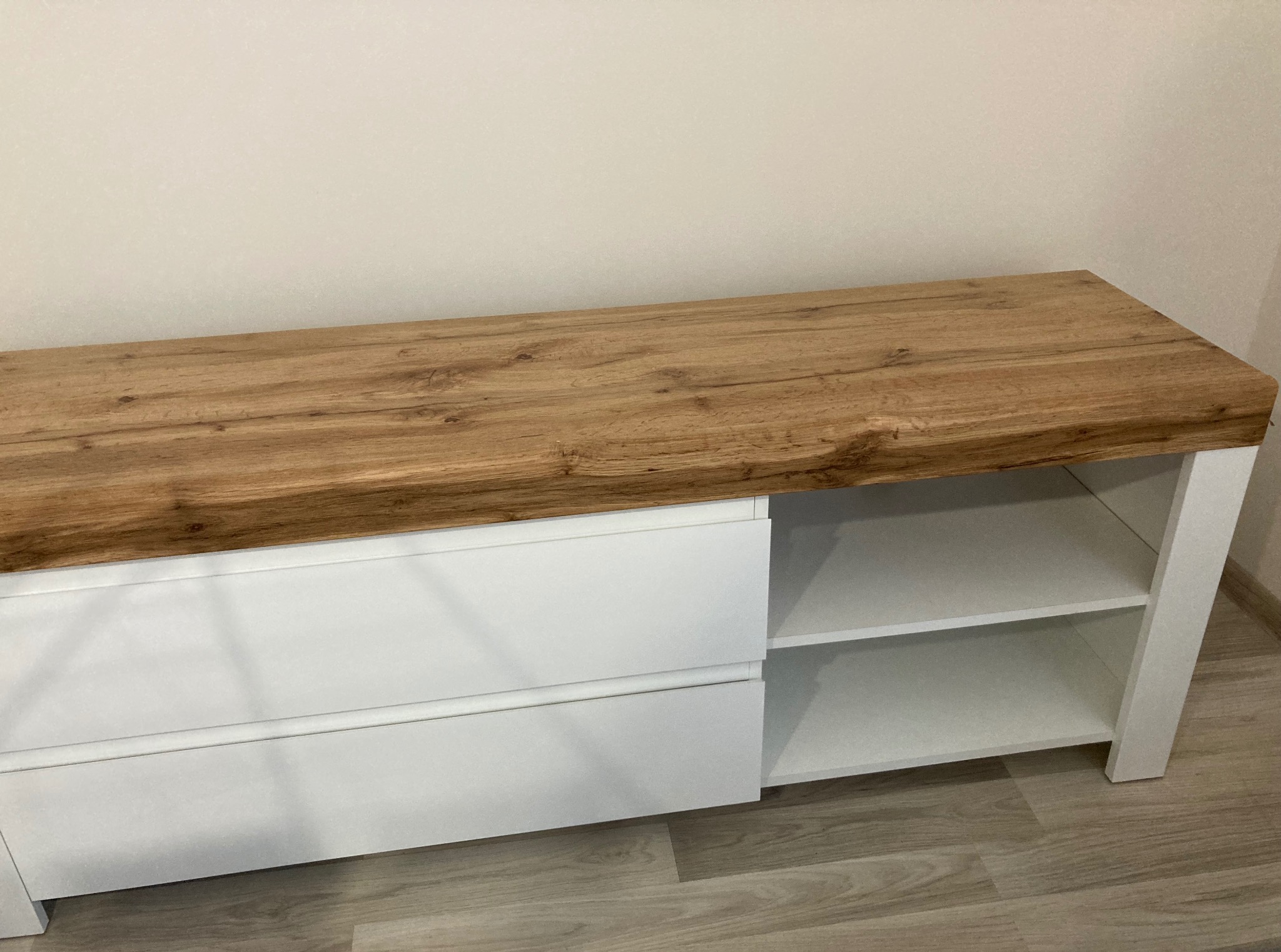 Nowoczesna komoda RTV z białymi frontami i blatem w odcieniu naturalnego drewna, na tle jasnej ściany i podłogi. Widoczne dwie szuflady i półki.