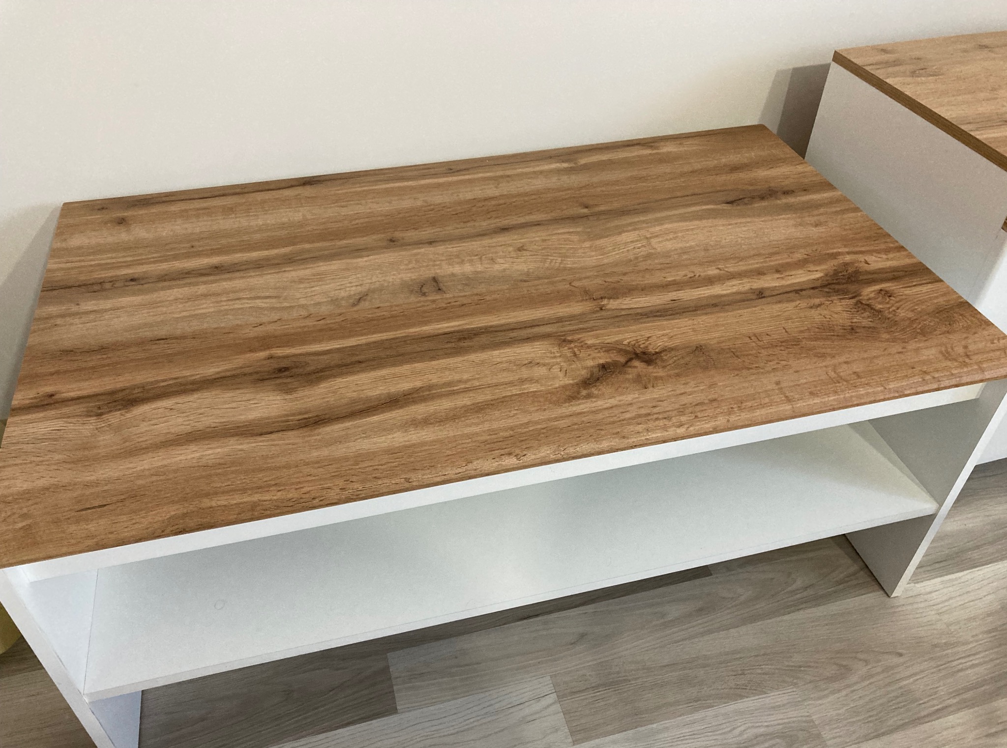 Zmontowany stolik kawowy z blatem imitującym drewno i białymi półkami, ustawiony na panelach podłogowych. Widoczny fragment drugiego mebla w tle.