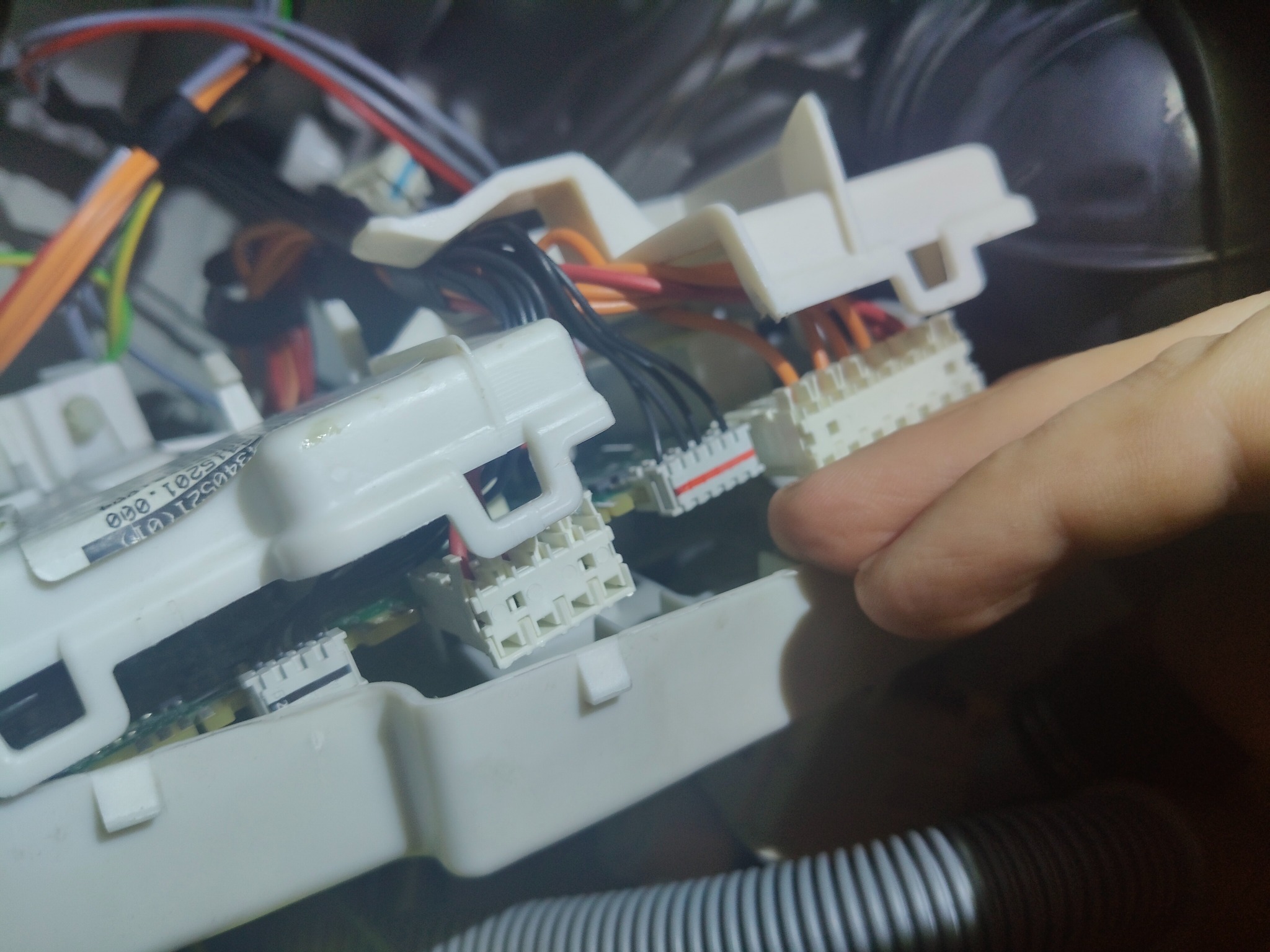 Wnętrze urządzenia AGD z widocznymi białymi modułami elektroniki, przewodami w kolorach: czarnym, pomarańczowym i czerwonym, oraz fragmentem dłoni w trakcie naprawy.