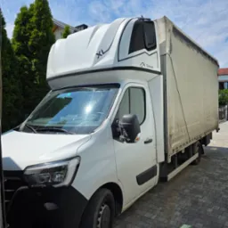 Biały bus dostawczy XL z plandeką marki Carpol na tle zielonych drzew i błękitnego nieba. Pojazd zaparkowany na brukowanej nawierzchni.