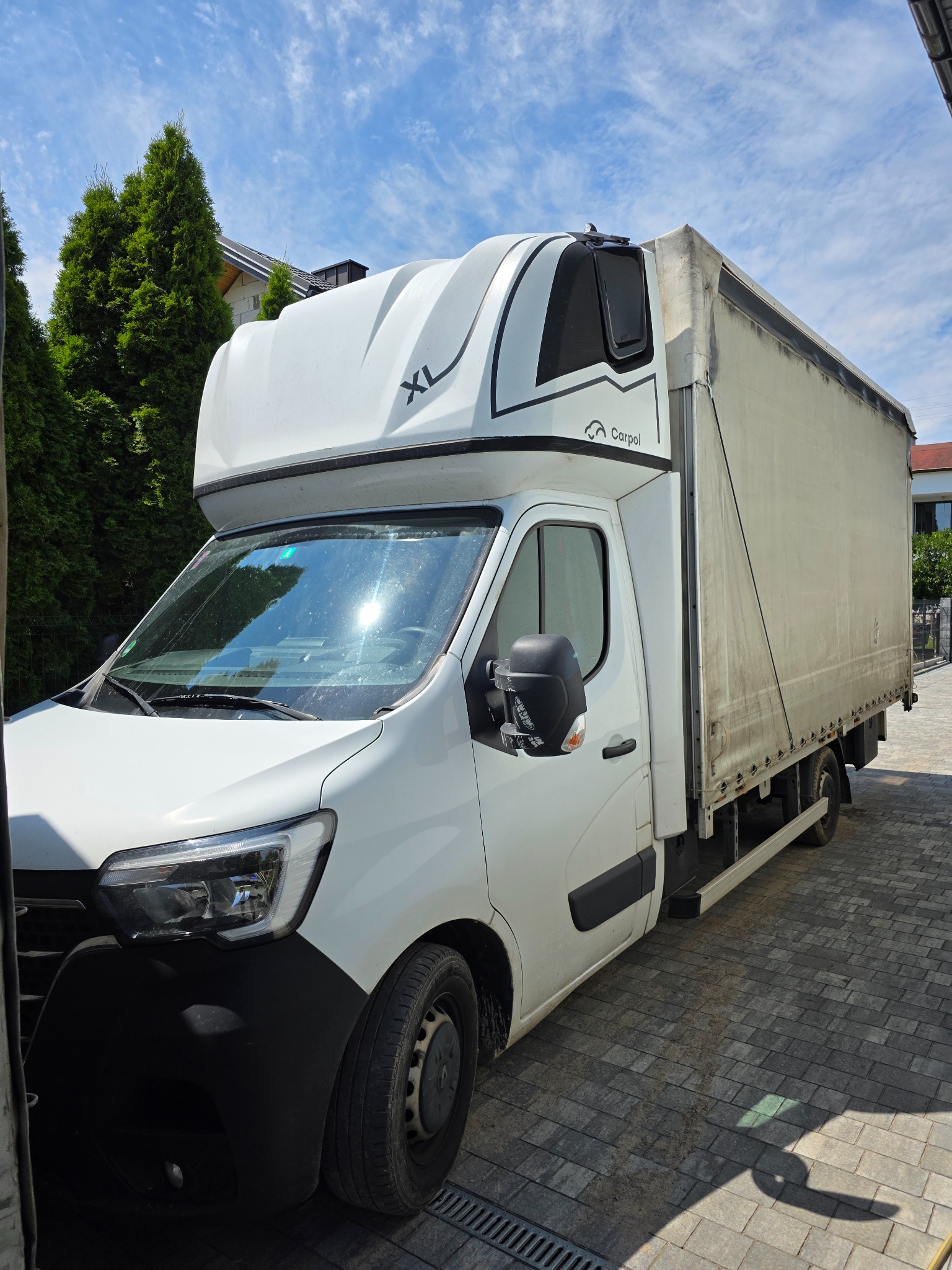 Biały bus dostawczy XL z plandeką marki Carpol na tle zielonych drzew i błękitnego nieba. Pojazd zaparkowany na brukowanej nawierzchni.