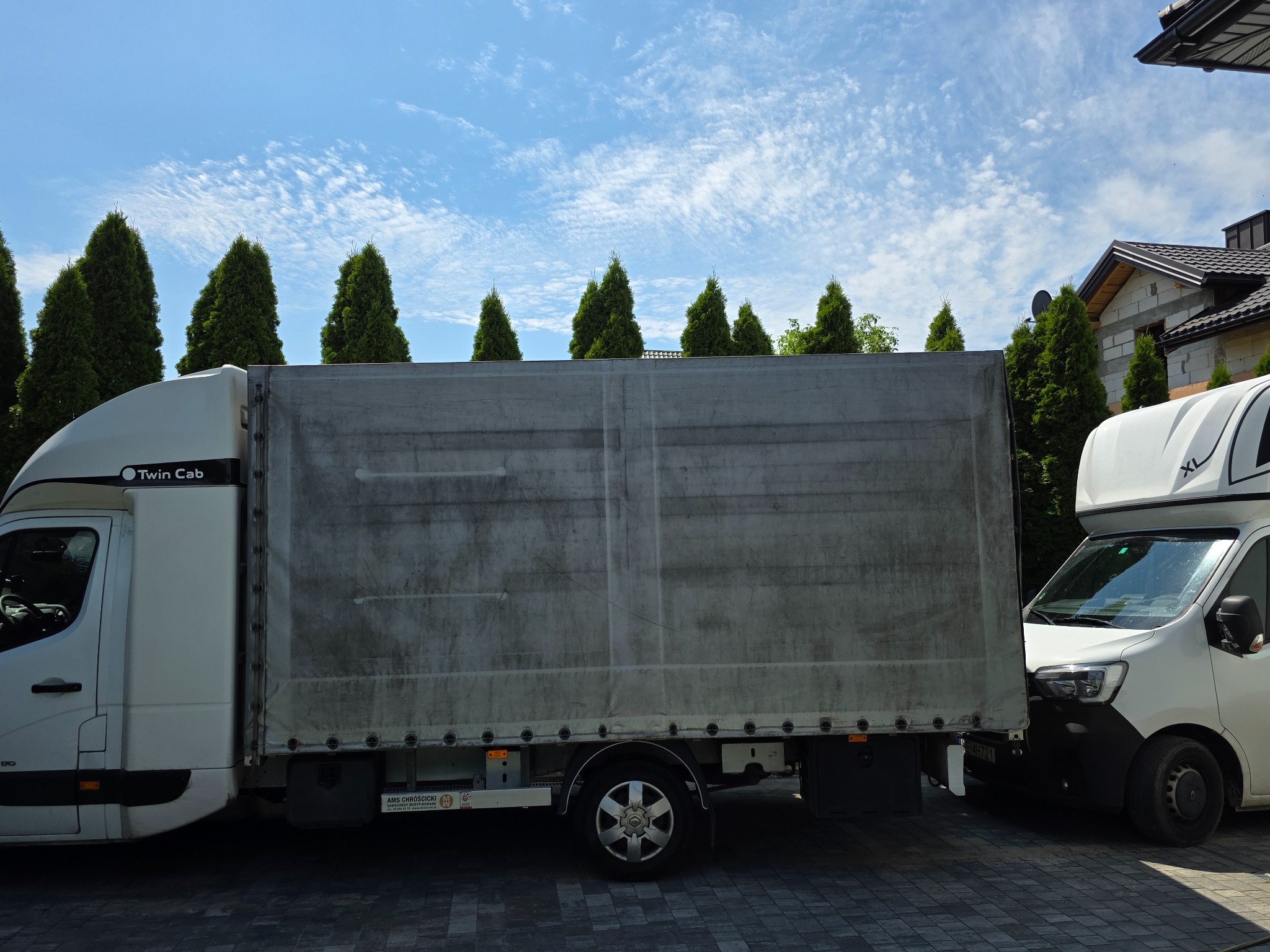 Dwa białe busy transportowe typu Twin Cab, zaparkowane na brukowanej nawierzchni, na tle iglaków i błękitnego nieba z obłokami.