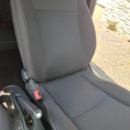 Czarne, tekstylne siedzenie samochodowe z jasnymi przeszyciami, widoczne elementy deski rozdzielczej i fragment otoczenia zewnętrznego.