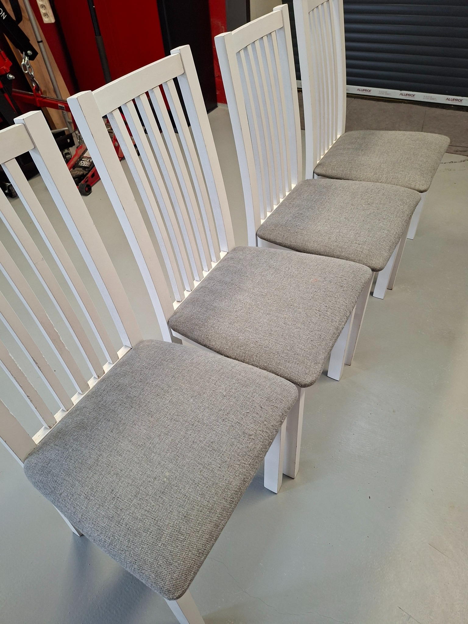 Cztery białe krzesła z szarą tapicerką ustawione w rzędzie na szarym tle. Widoczne elementy warsztatowe w tle.