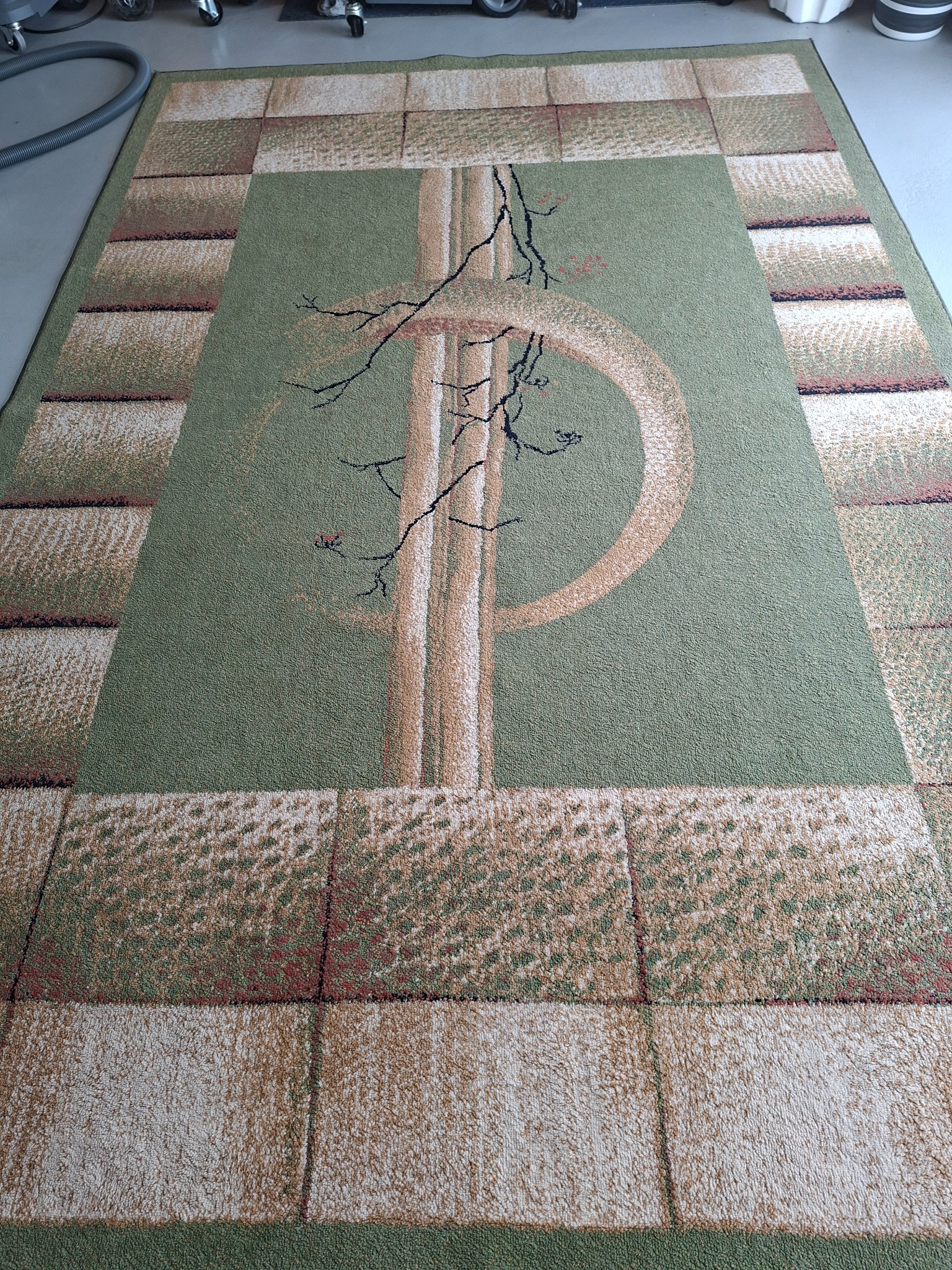 Wyczyszczony, zielony dywan z geometrycznym wzorem i motywem drzewa, leżący na szarej podłodze. Widoczny wąż od odkurzacza w tle po lewej stronie.