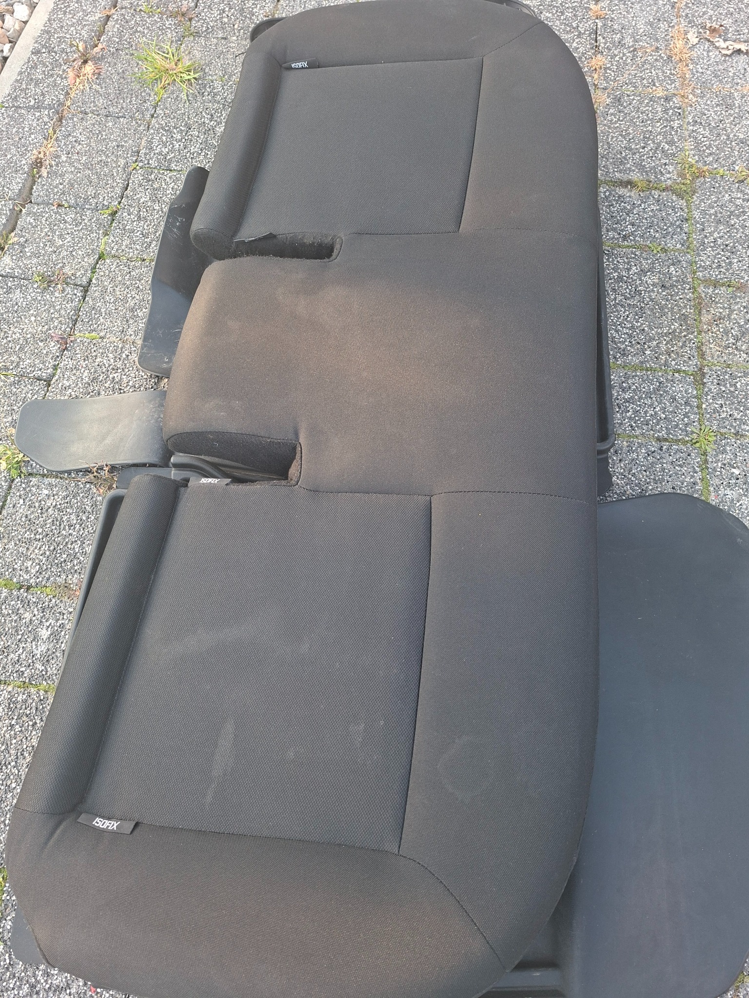 Ciemna, materiałowa tapicerka samochodowa z widocznymi elementami mocującymi ISOFIX, leżąca na szarej kostce brukowej z fragmentami roślinności.