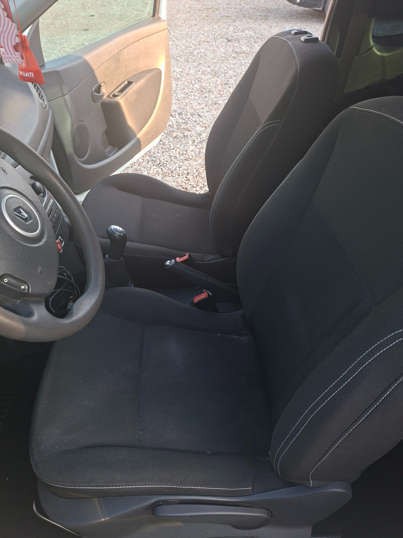 Wnętrze samochodu z czarnymi fotelami, widoczne ślady użytkowania na tapicerce. Otwarta biała klapa drzwi, kierownica i dźwignia zmiany biegów.