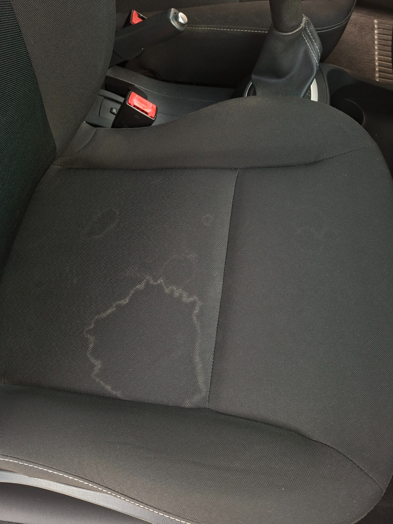 Czarny fotel samochodowy z wyraźną plamą na siedzisku. Widoczny fragment pasów bezpieczeństwa i dźwigni zmiany biegów. Tapicerka tekstylna, wymagająca czyszczenia.