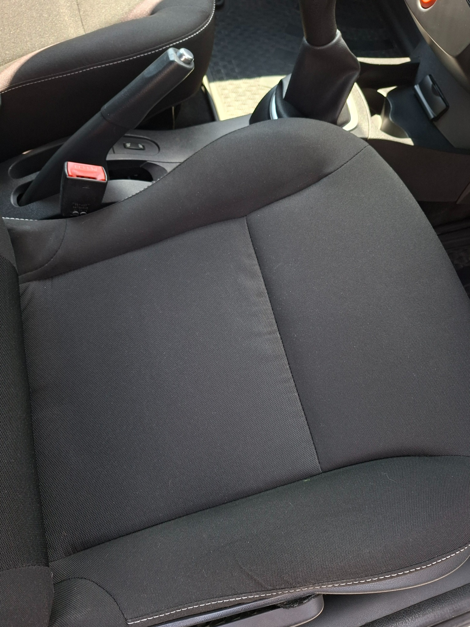 Czarne, tekstylne siedzenie samochodowe z widocznym hamulcem ręcznym i dźwignią zmiany biegów. Detal wnętrza pojazdu w zbliżeniu, stonowana kolorystyka.