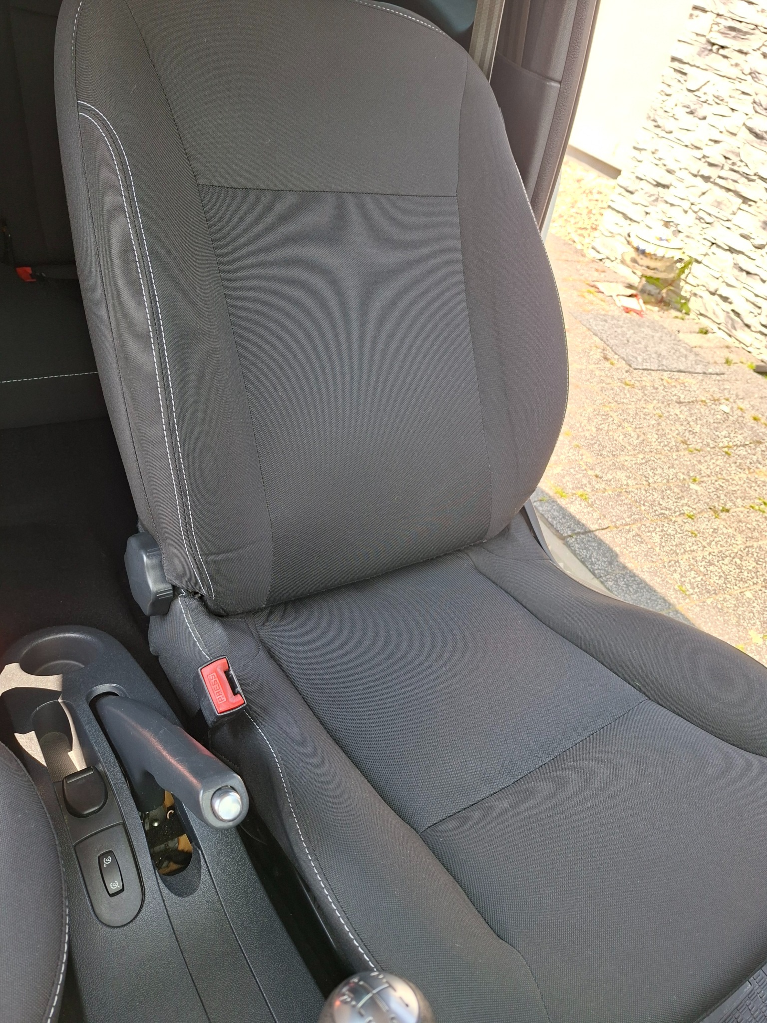 Czarne, tekstylne siedzenie samochodowe z jasnymi przeszyciami, widoczne elementy deski rozdzielczej i fragment otoczenia zewnętrznego.