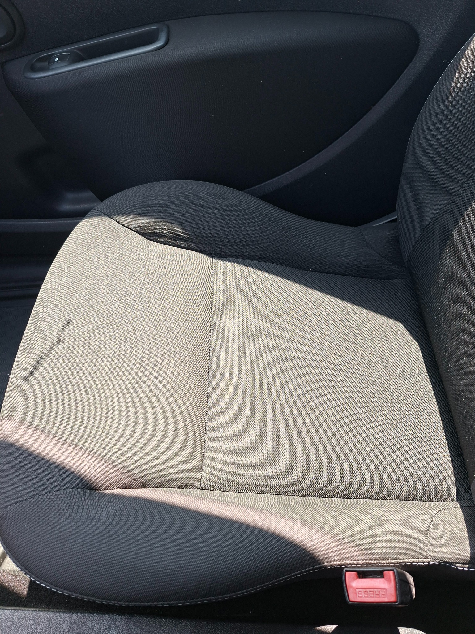 Szare, materiałowe siedzenie samochodowe z widocznym zapięciem pasa bezpieczeństwa i fragmentem drzwi. Widoczne ślady użytkowania.